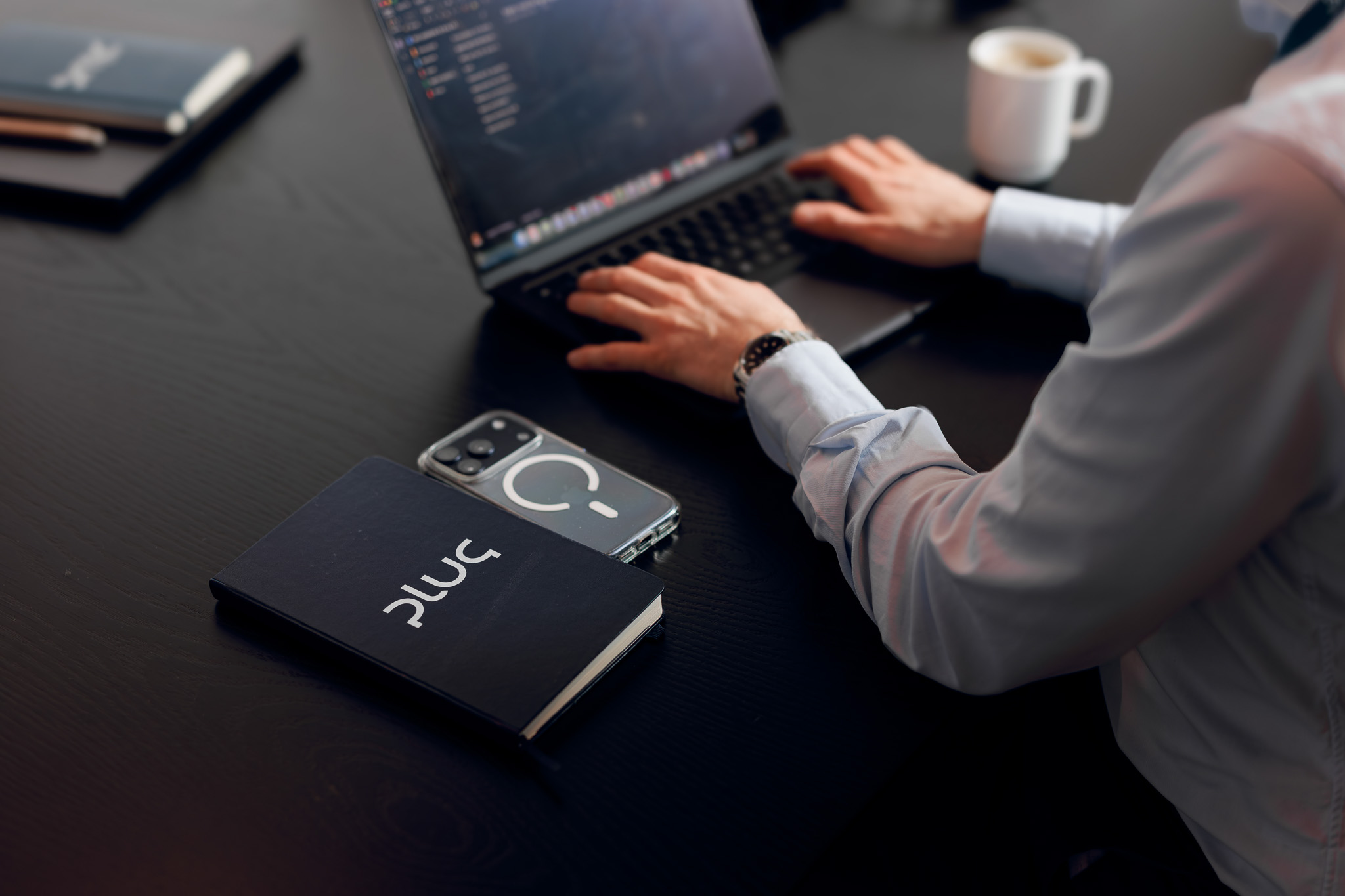Person typing on a laptop at a desk with a black notebook labelled 'PLUG' and a smartphone with a circular design case beside it.
