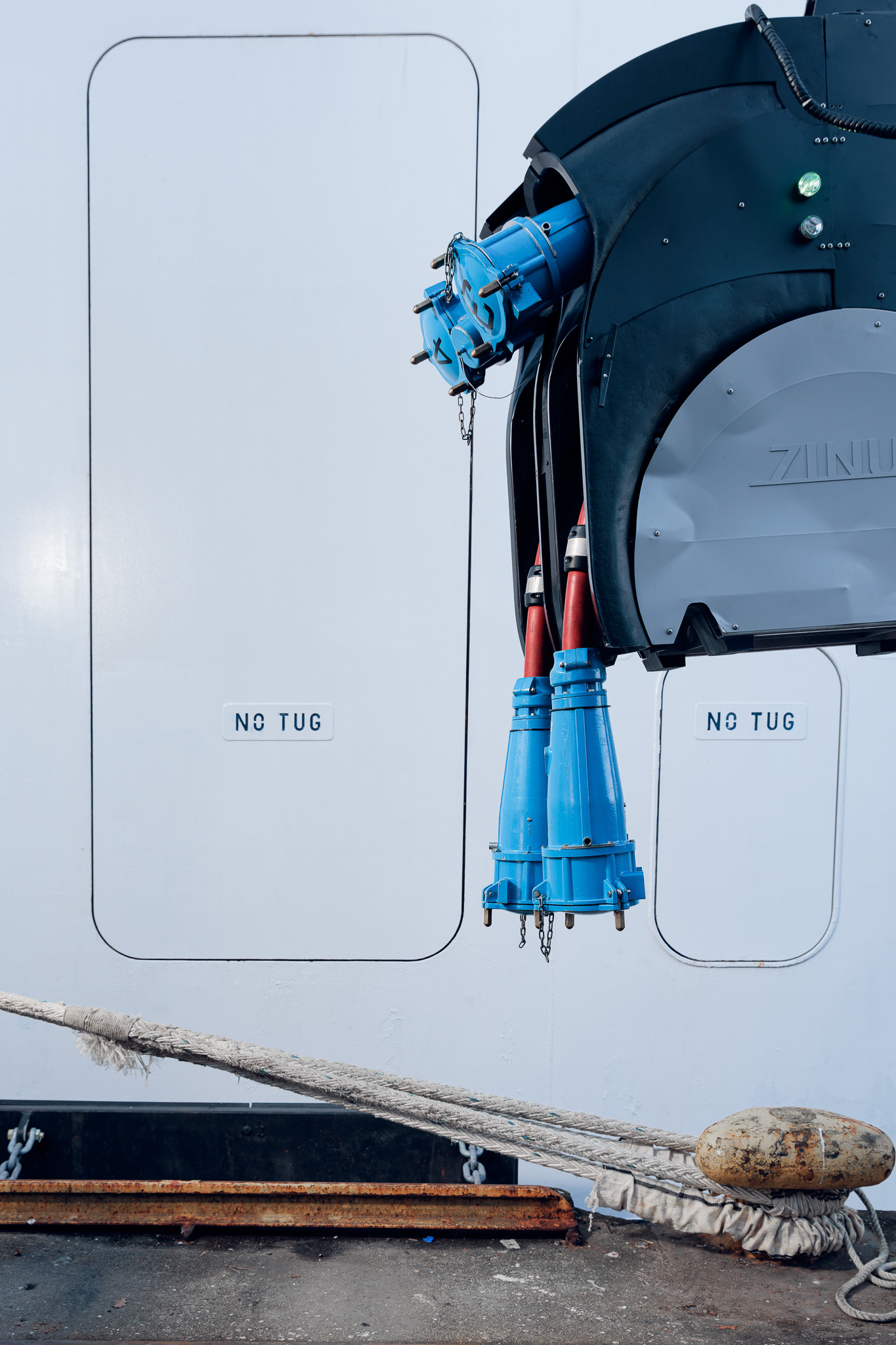 Large blue ship electrical connectors hanging near white ship wall with 'NO TUG' signs and coiled ropes on dock.
