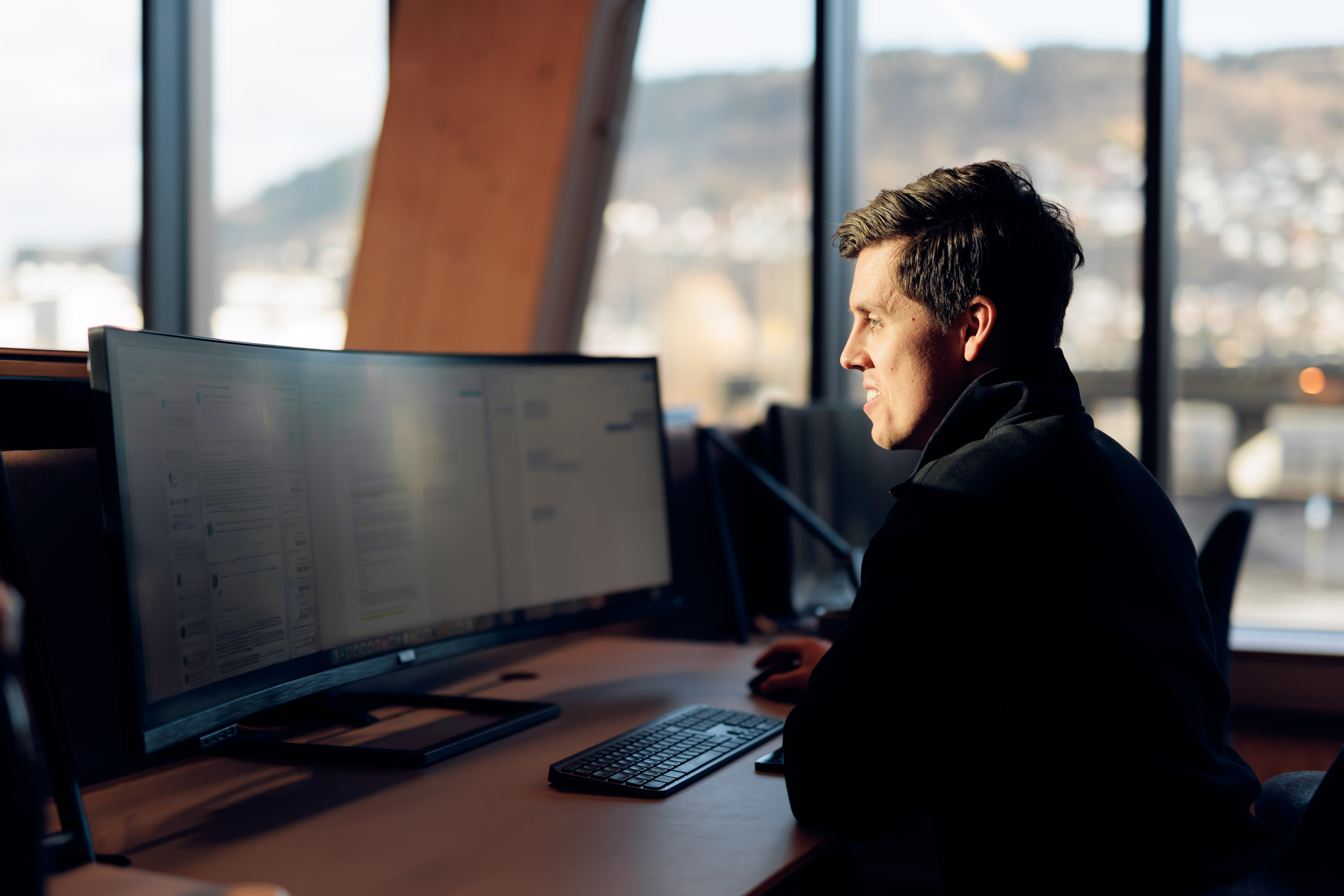 Young man working on a computer with an ultra-wide curved monitor in an office with large windows and a mountain view.