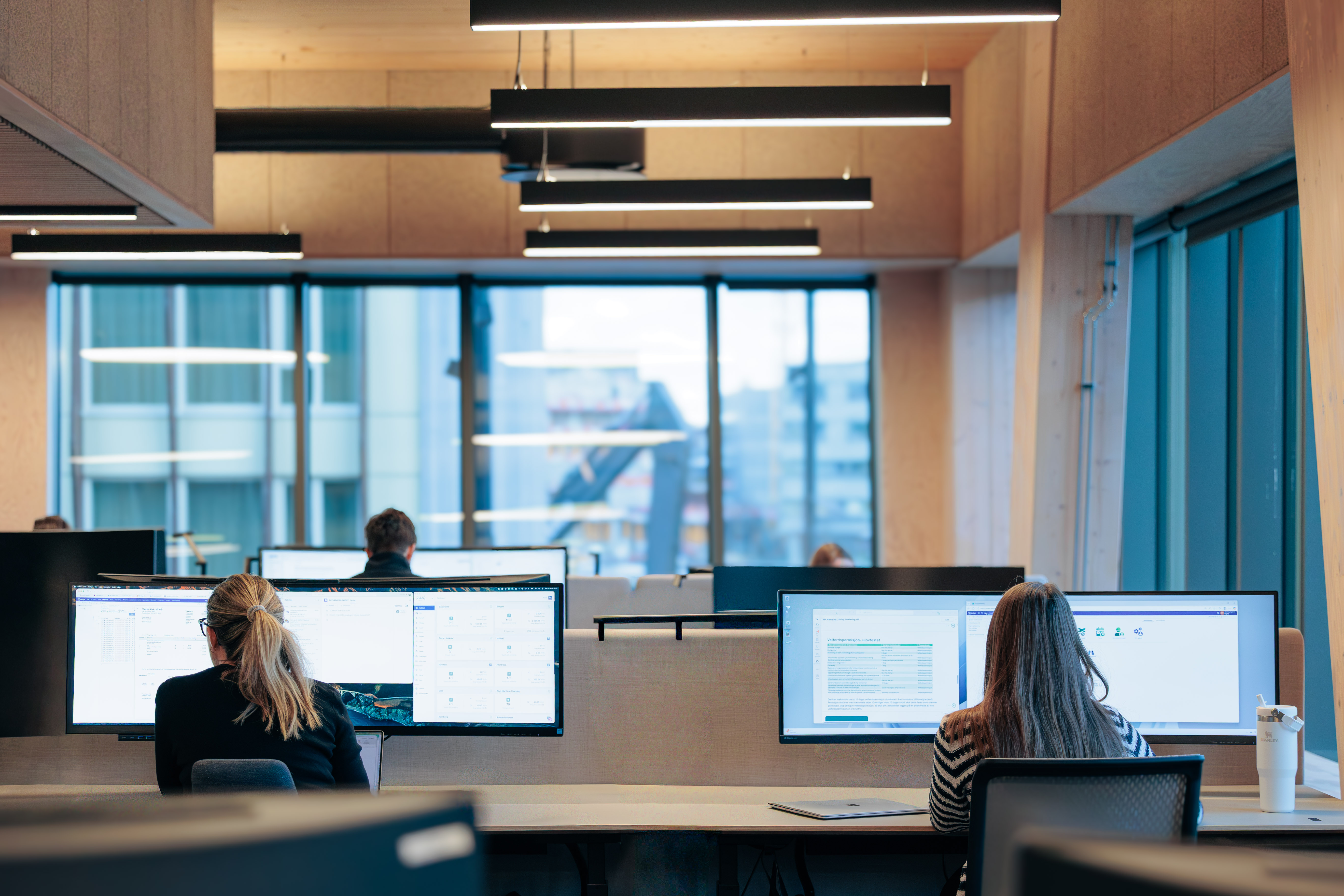 Two women working on computers with large monitors in a modern open office space with wooden walls and large windows.