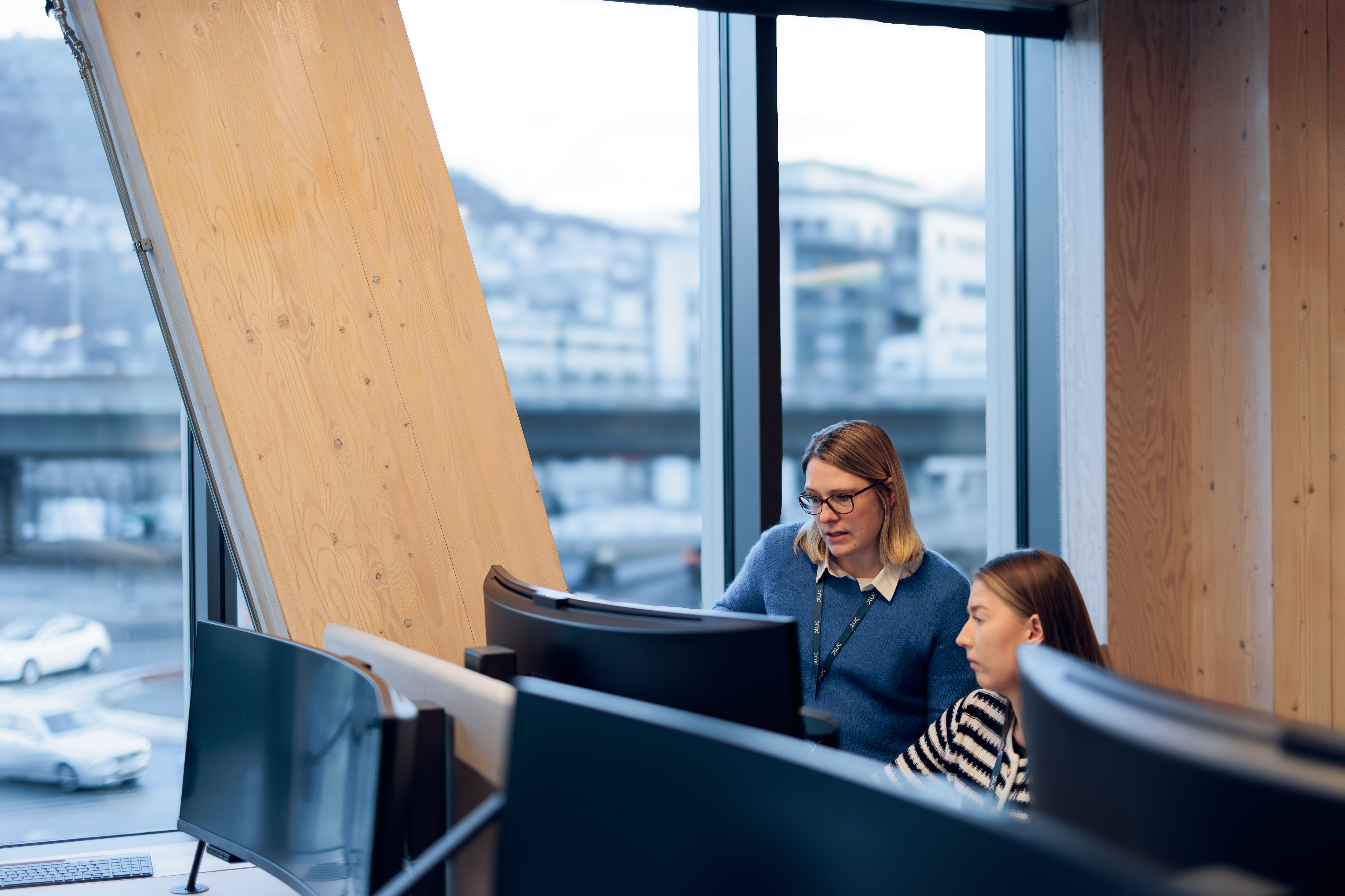 Two women working together at computer monitors in a modern office with large windows and wooden paneling.