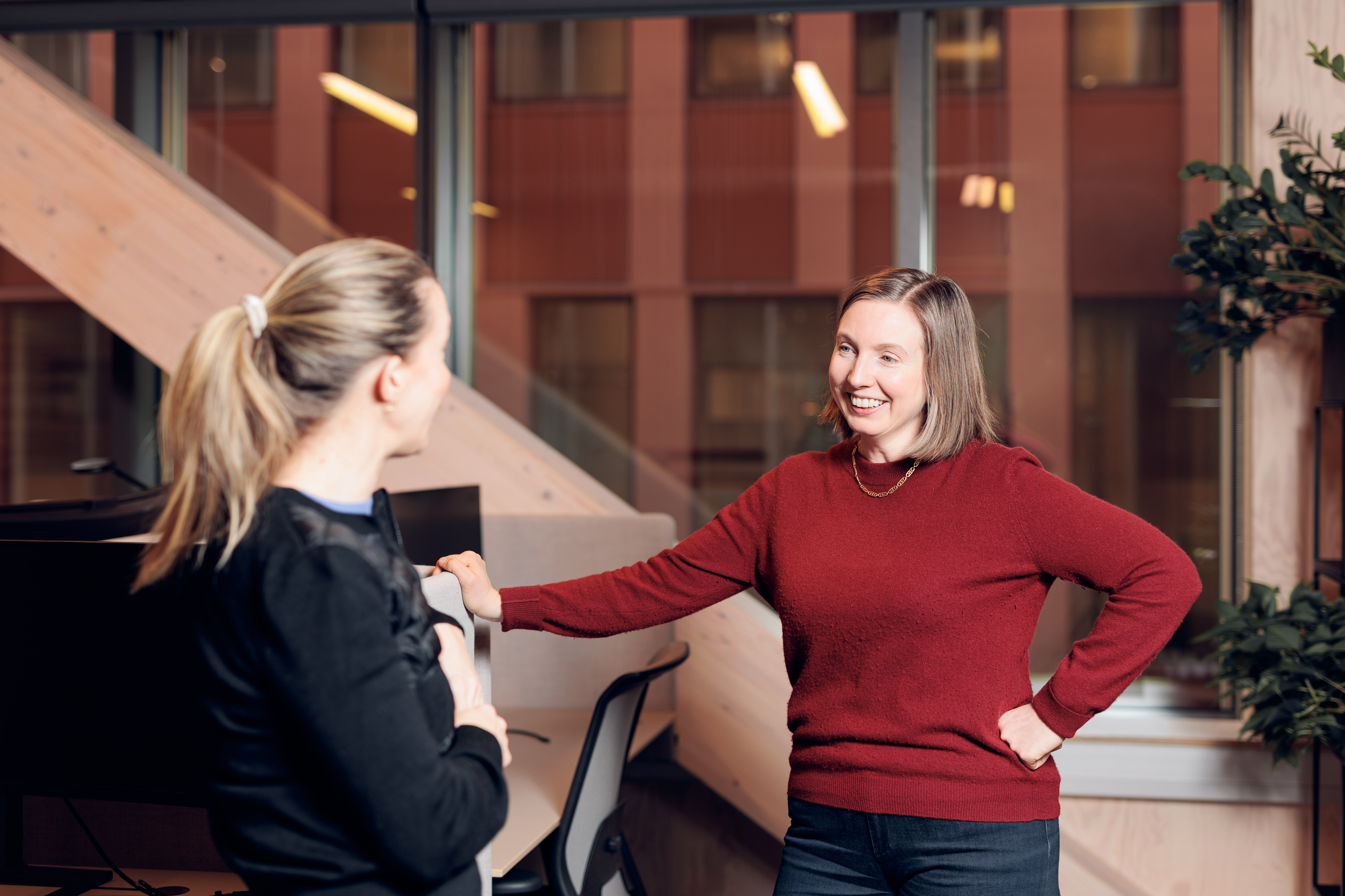 Two women chatting in a modern office, one wearing a red sweater and smiling, the other with a ponytail in a black top.