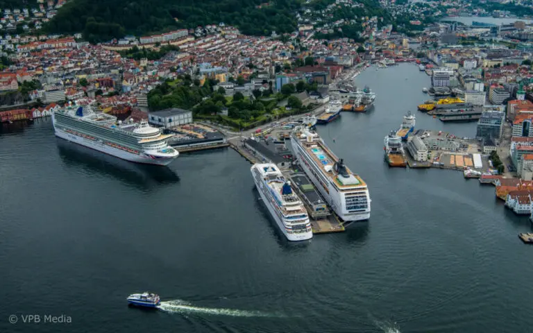 Aerial view of a harbor with three large cruise ships docked and a small boat moving in the water, surrounded by a city with buildings and green hills.