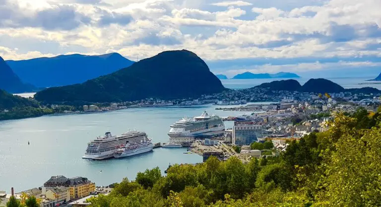 Two large cruise ships docked in a coastal town surrounded by green hills and mountains under a partly cloudy sky.