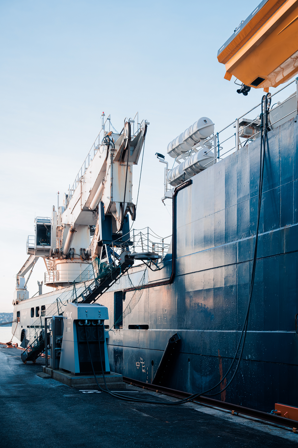 Large docked ship with white cranes and lifeboats on the deck during daylight.