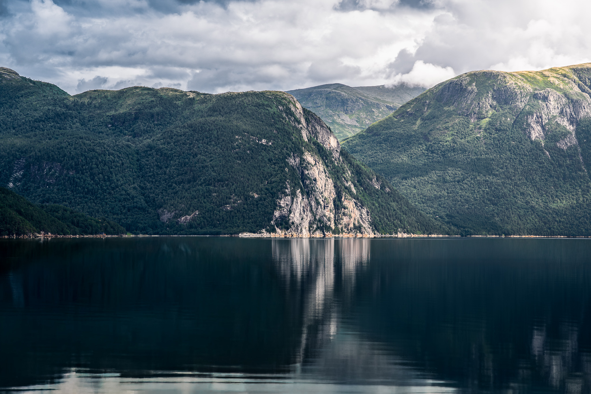 Calm lake reflecting forested mountains under a cloudy sky.