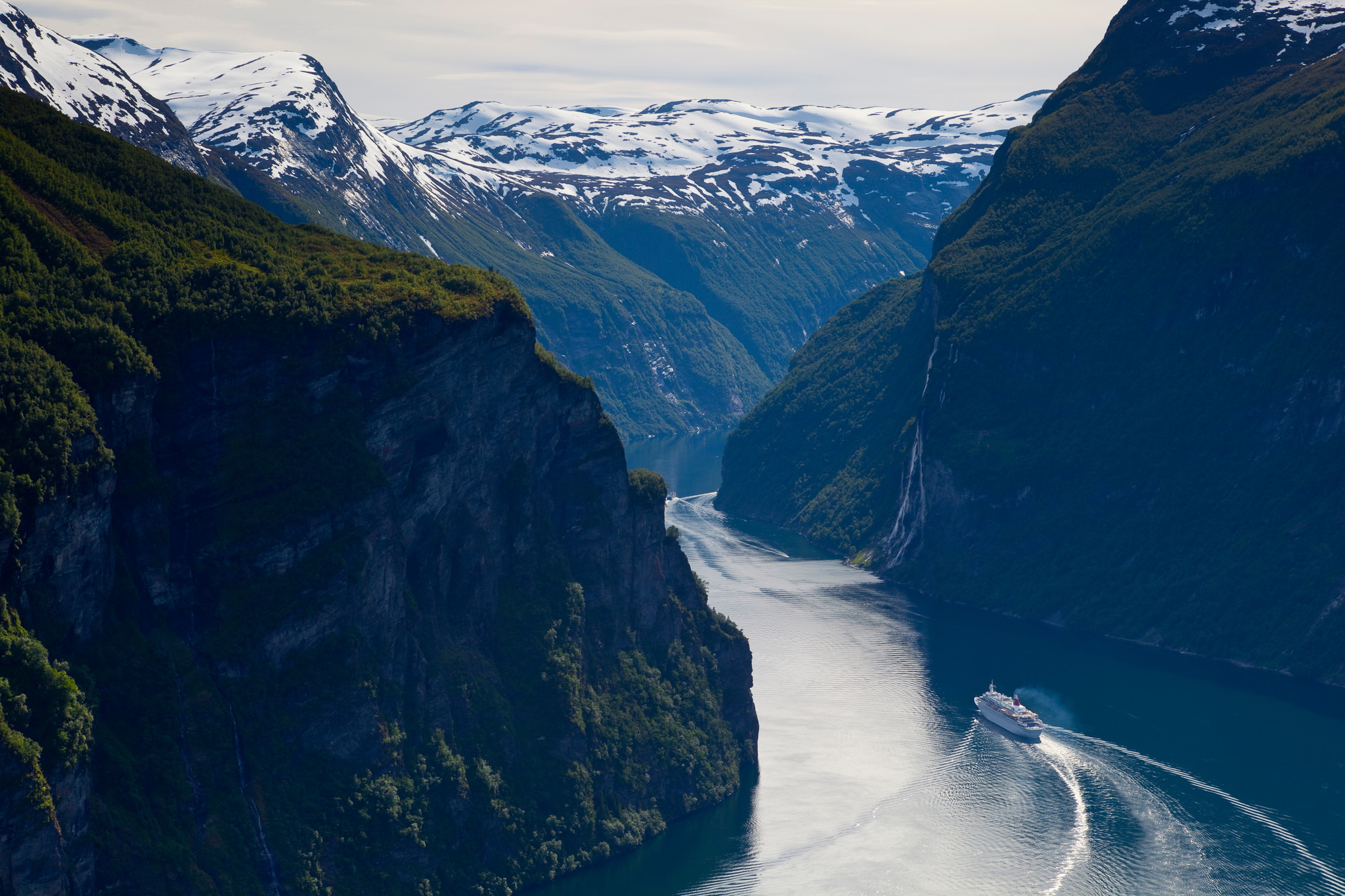 Cruise ship sailing through a deep fjord with steep green cliffs and snow-capped mountains in the background.