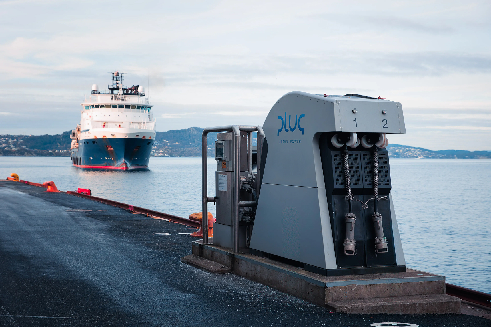 Dockside shore power unit labeled 'Plug' with a large ship approaching in the background over calm water.