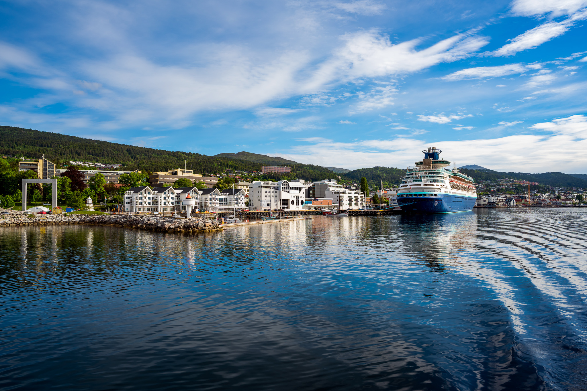 Large cruise ship docked at a waterfront town with modern buildings and forested hills under a blue sky with scattered clouds.