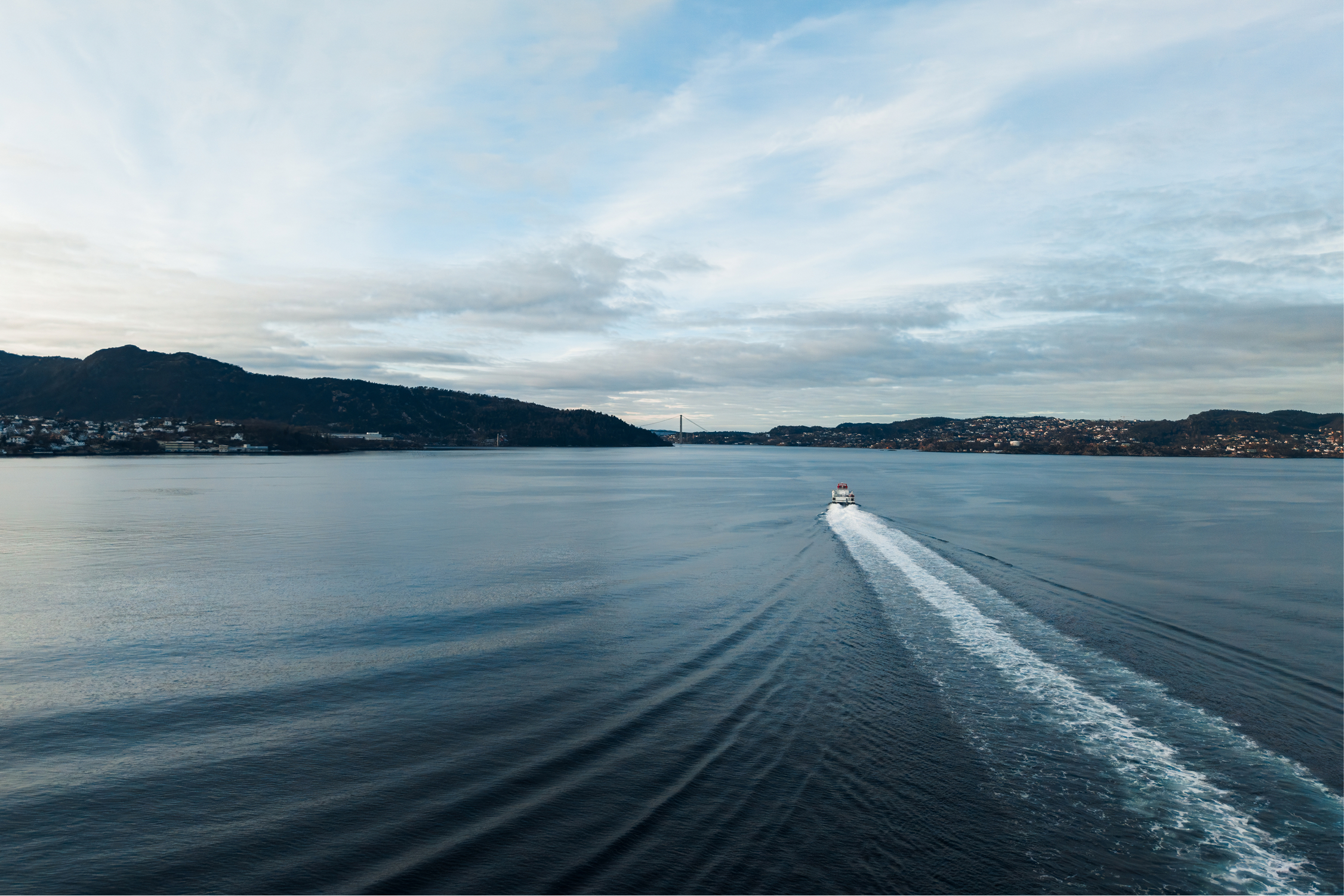 Small boat creating a white wake as it moves through calm water with hills and a bridge in the distance under a cloudy sky.