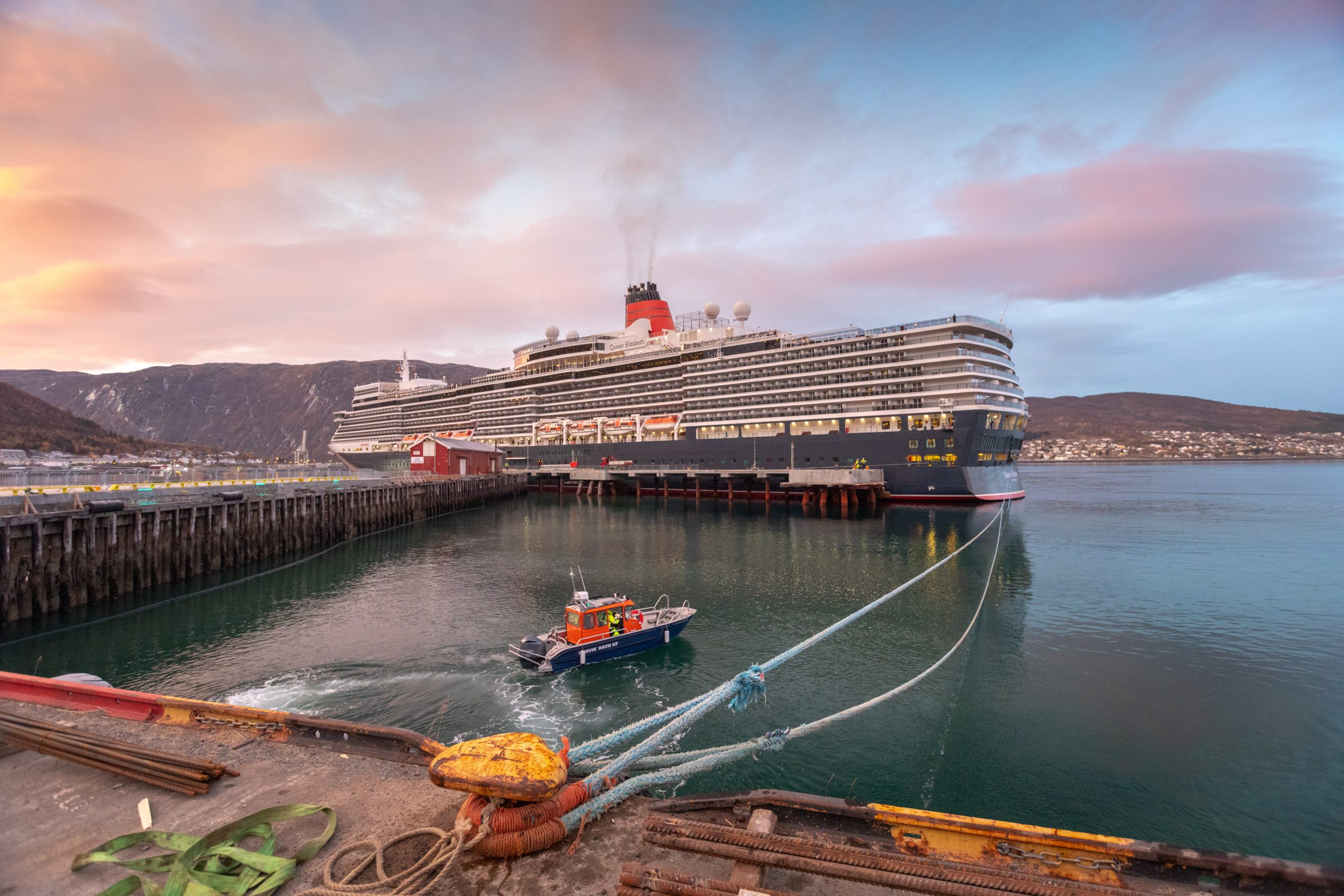 Large cruise ship Queen Elizabeth docked at a harbor with a small boat nearby and mountains in the background during sunset.
