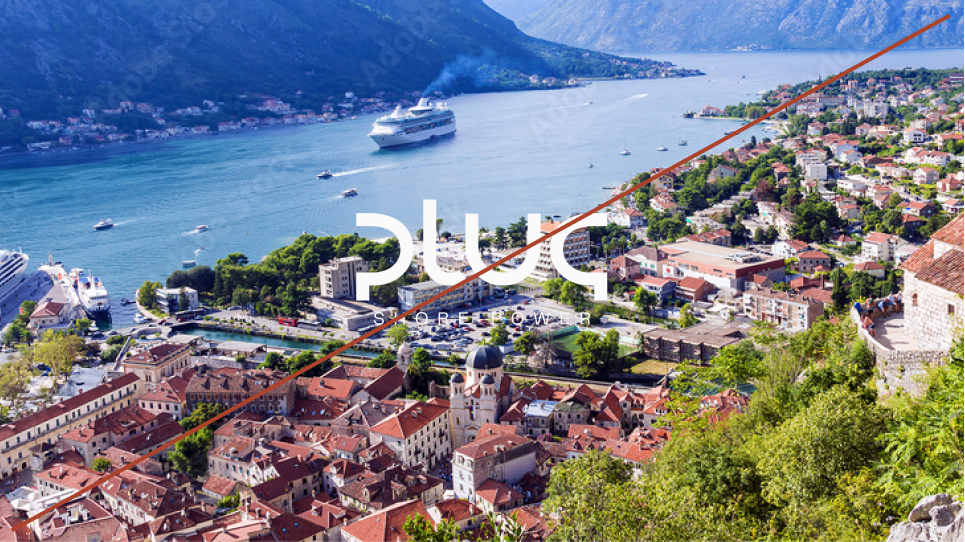 Aerial view of a coastal town with red-roofed buildings near a fjord with boats and cruise ships in the water, surrounded by mountains.