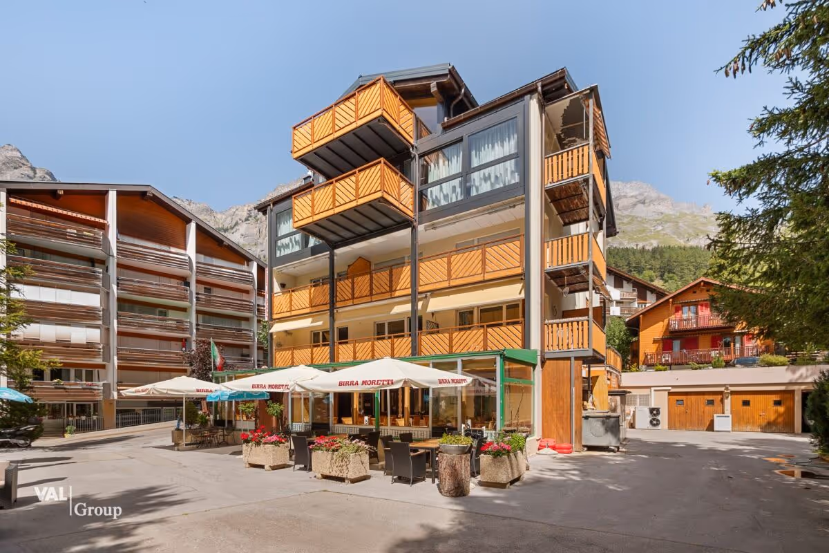 Dreistöckiges Gebäude mit Holzbalustraden und Außenterrasse mit Sonnenschirmen vor Bergkulisse.