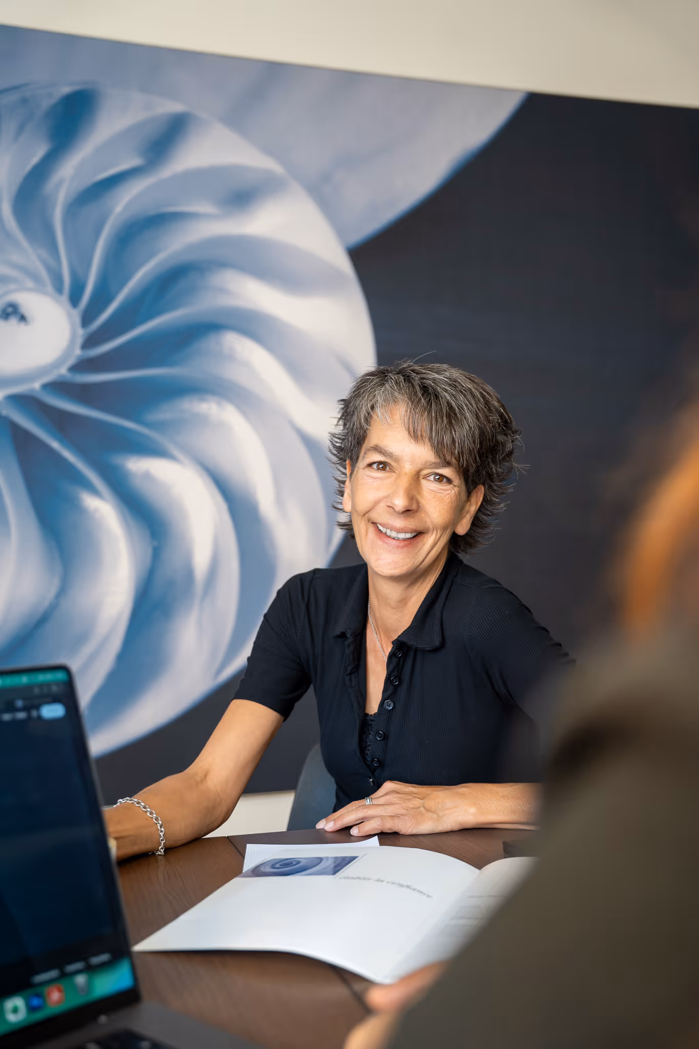 Frau mit kurzem grauem Haar, die an einem Tisch mit geöffnetem Buch lächelt, vor einer blauen abstrakten Wandkunst.