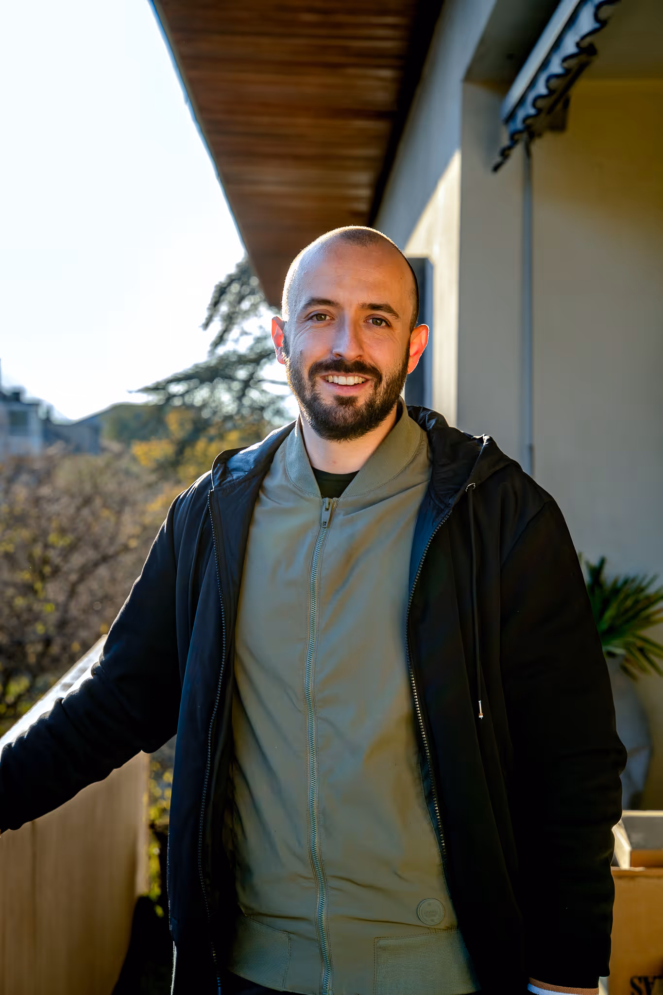 Mann mit Bart und Glatze lehnt lächelnd an einem Balkon mit Bäumen im Hintergrund.