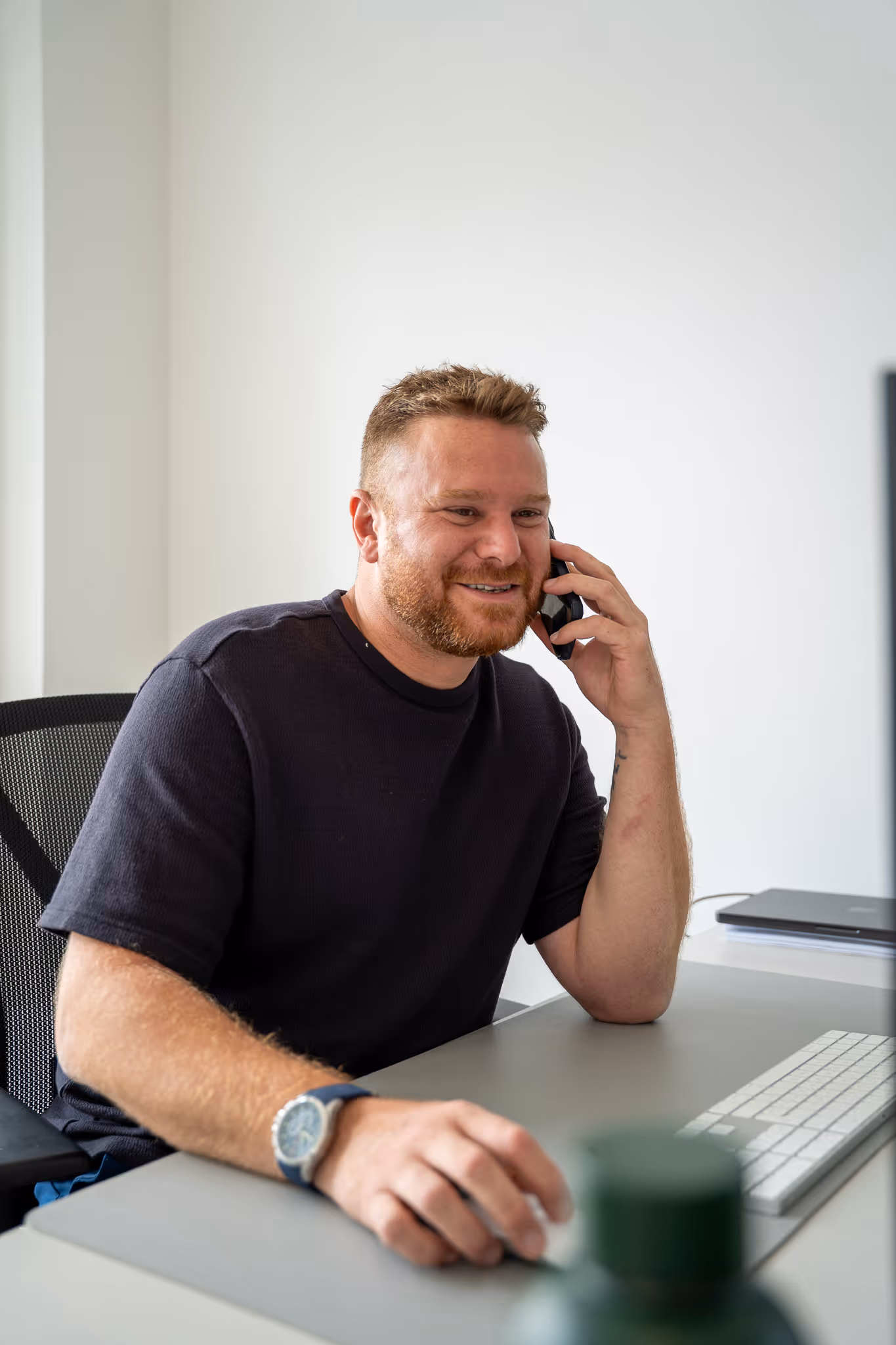 Lächelnder Mann mit kurzem Haar und Bart telefoniert am Schreibtisch mit Computer und Tastatur.