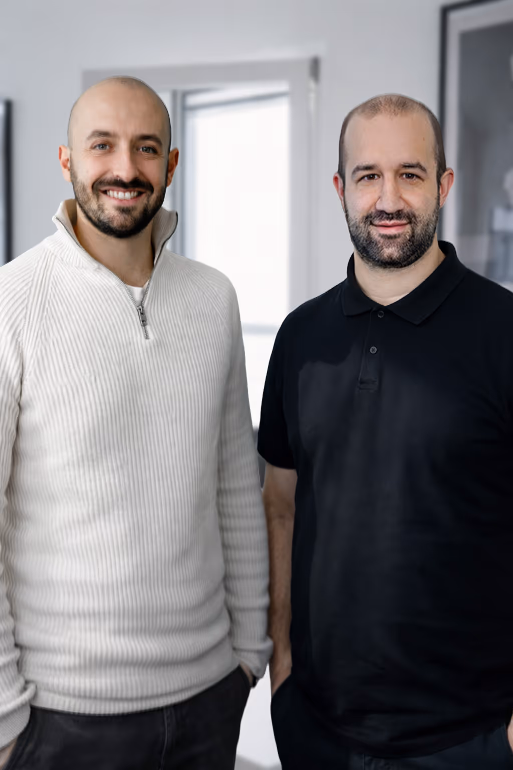 Zwei lächelnde Männer, einer in einem weißen Pullover mit Reißverschluss, der andere in einem schwarzen Poloshirt, stehen nebeneinander in einem hellen Raum.