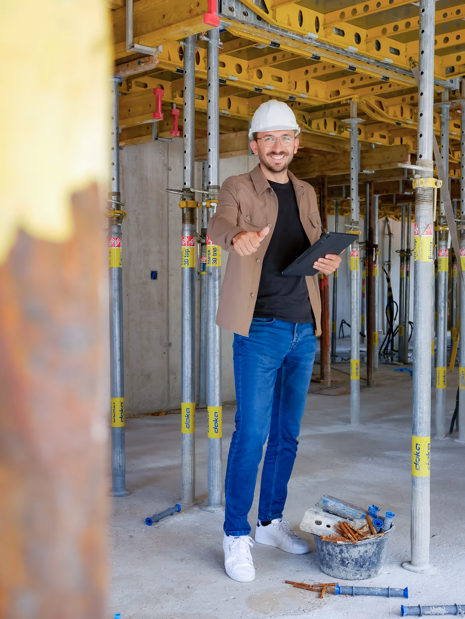 Ein Bauleiter mit Schutzhelm hält ein Tablet und zeigt auf einer Baustelle mit Metallstützen und Bauschutt den Daumen nach oben.