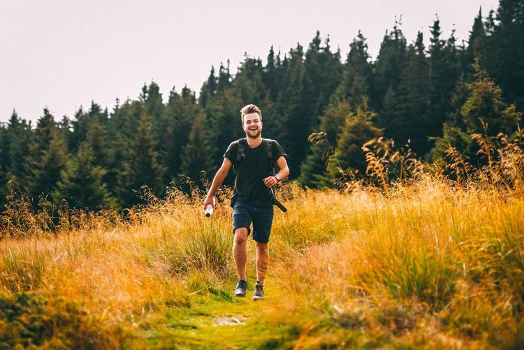 Smiling man with a backpack running on a grassy path surrounded by tall golden grass and pine trees in the background.
