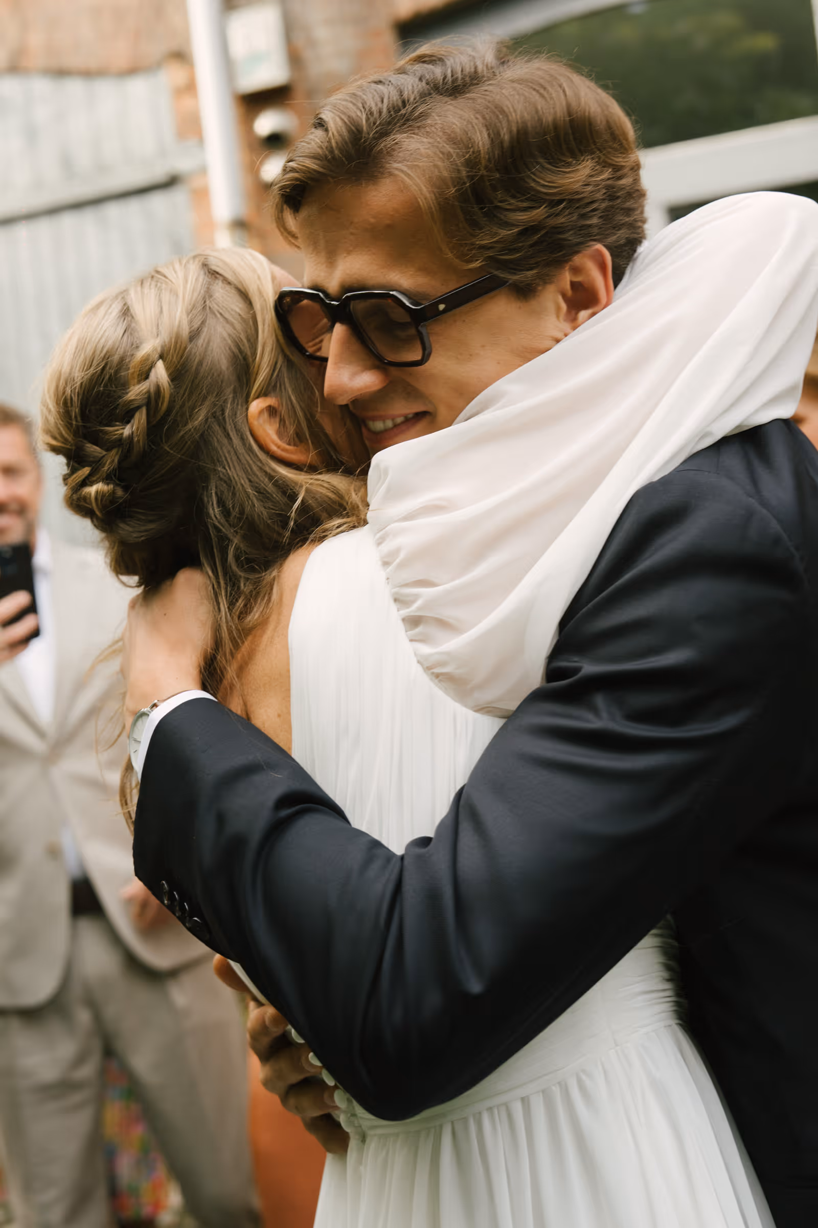 Man in glasses and dark suit hugging a woman in a white dress with braided hair at an outdoor event.