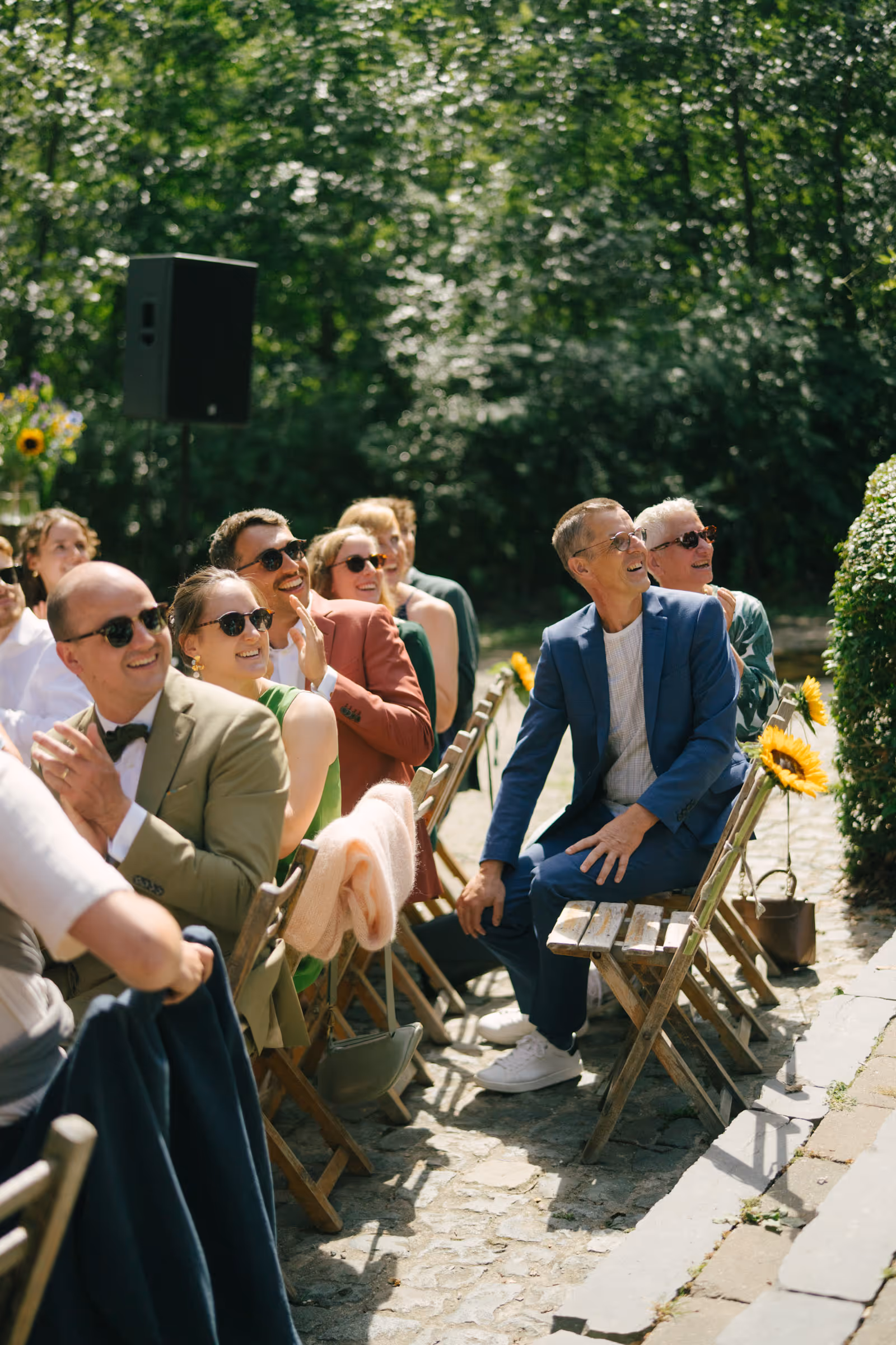 Group of smiling people wearing sunglasses sitting on wooden chairs outdoors, looking and clapping towards the right.