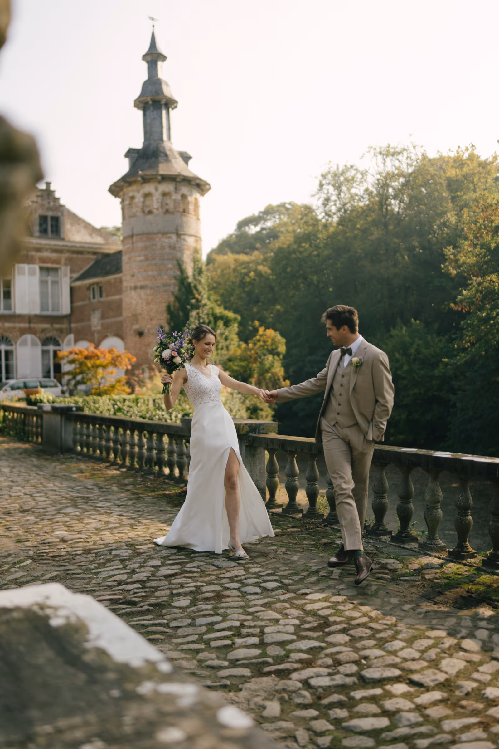 Bride in white dress holding a bouquet and groom in beige suit holding hands on a cobblestone path near a historic stone building and greenery.