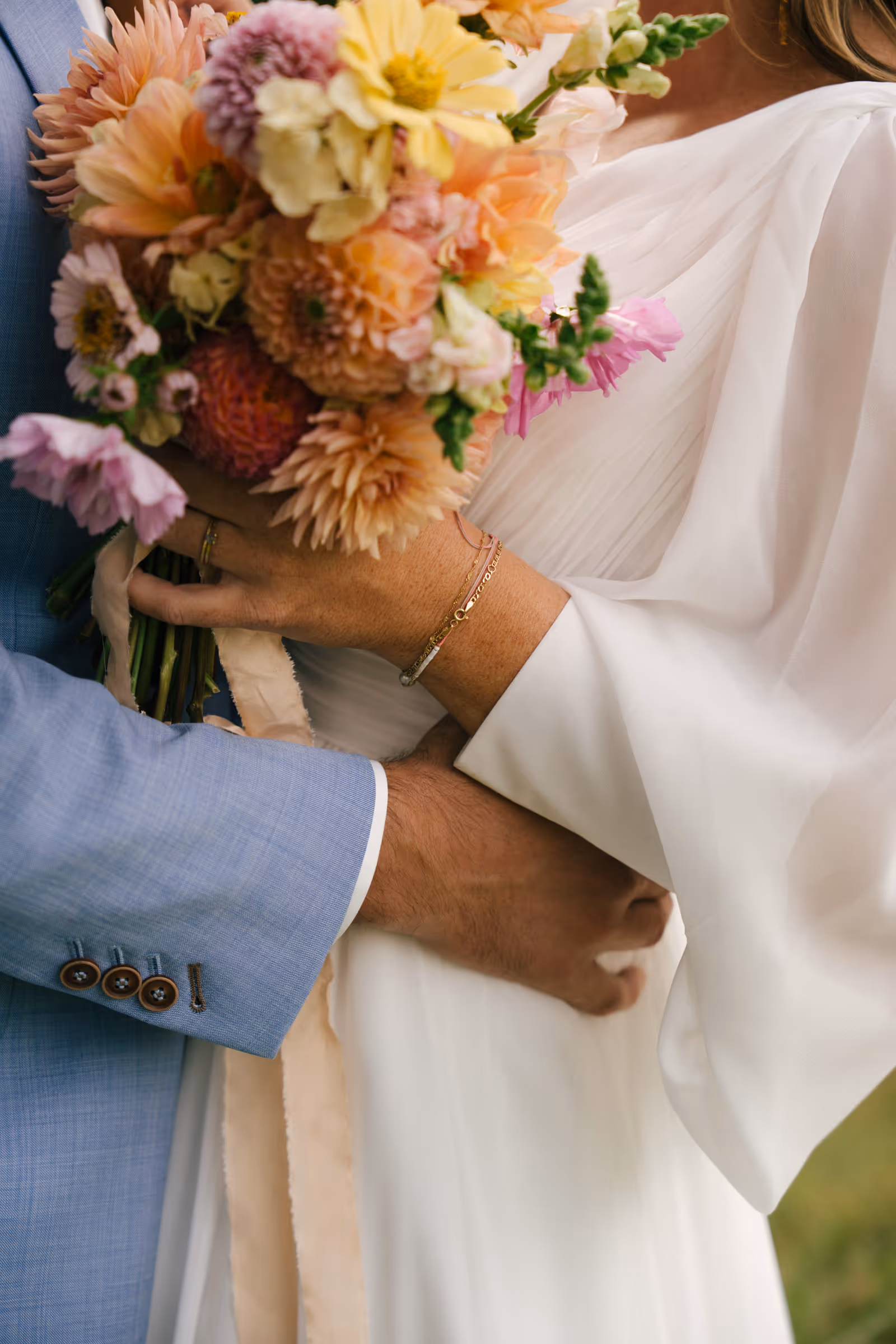 Close-up of a bride and groom holding a colorful bouquet with the groom in a light blue suit and the bride in a white dress.