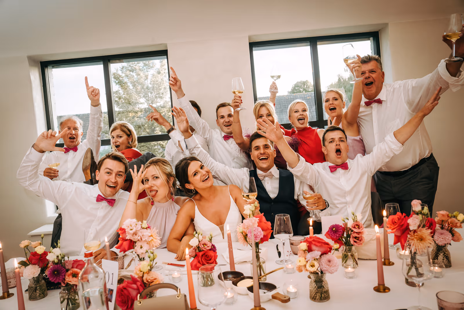 A joyful group of wedding guests raising glasses and cheering around a table with flowers and candles.