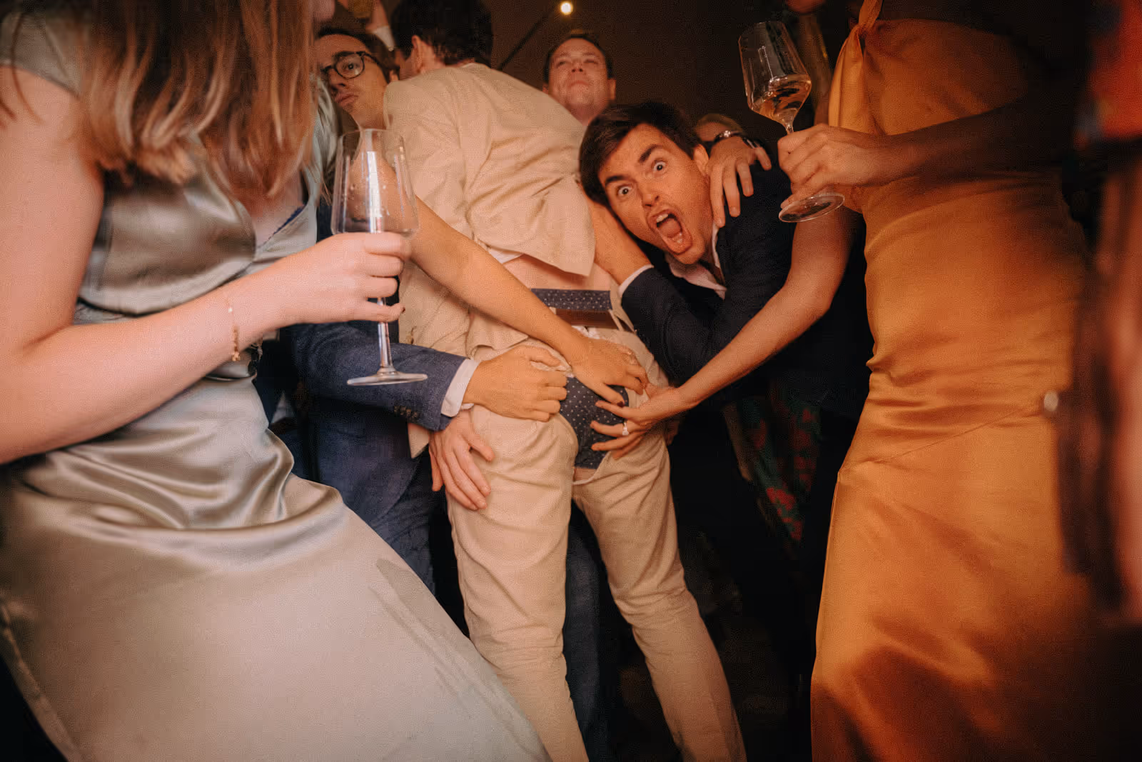 Group of people at a party, one person bending over showing polka-dot underwear while others touch and pose around them with drinks in hand.