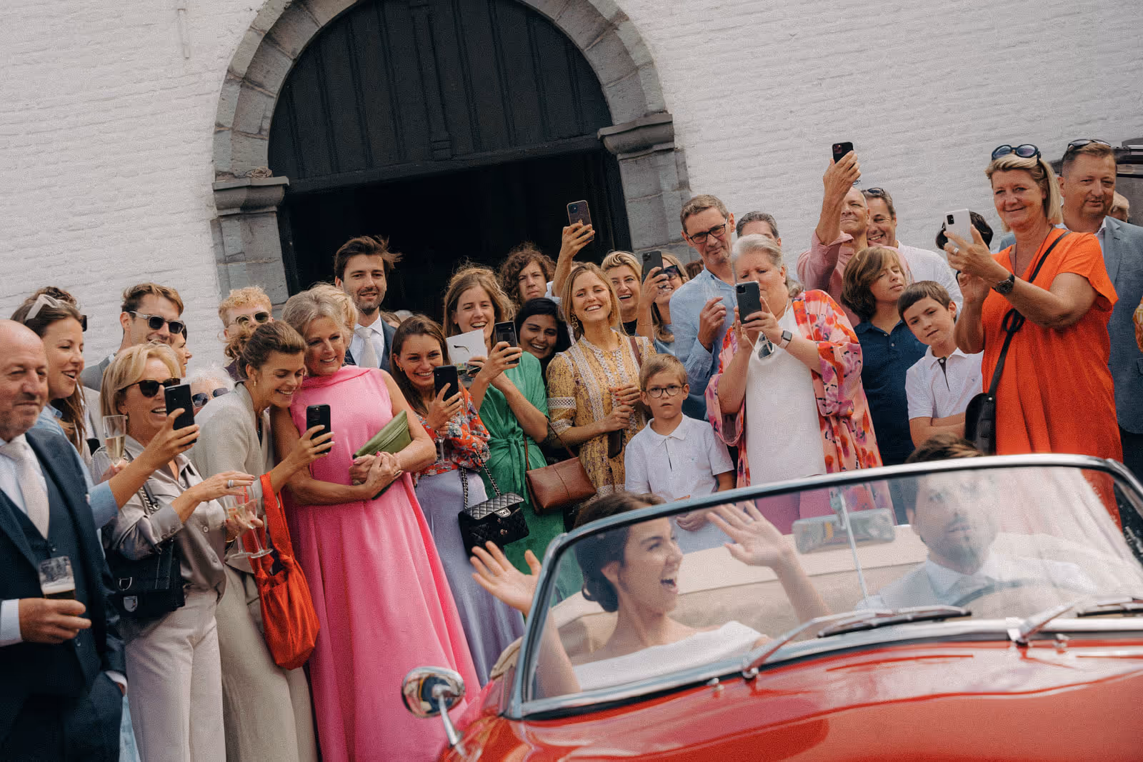 Group of people happily taking photos of a woman in a white dress and a man driving a red convertible car outside a building with an arched doorway.