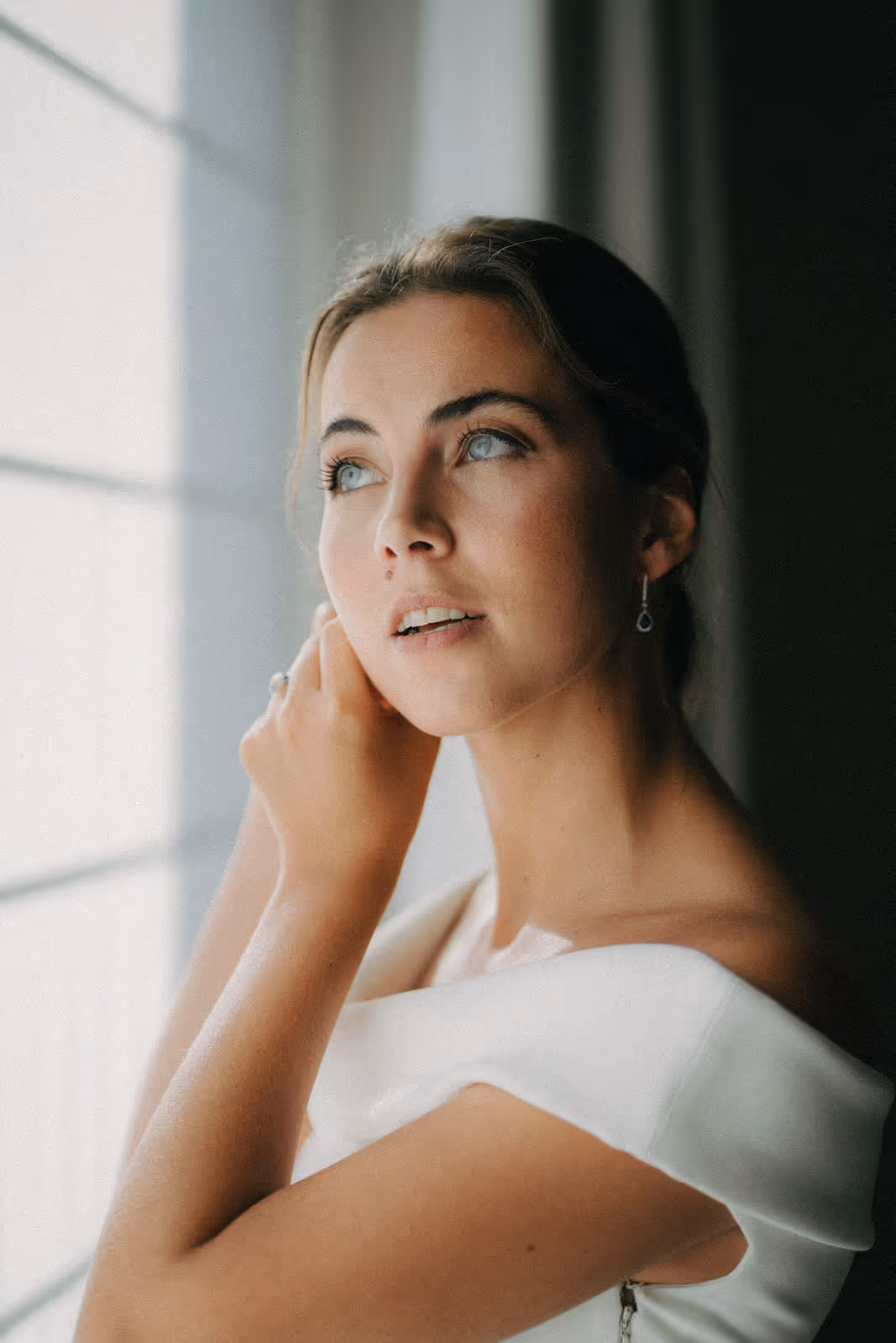 Woman in a white dress looking out a window while adjusting an earring.
