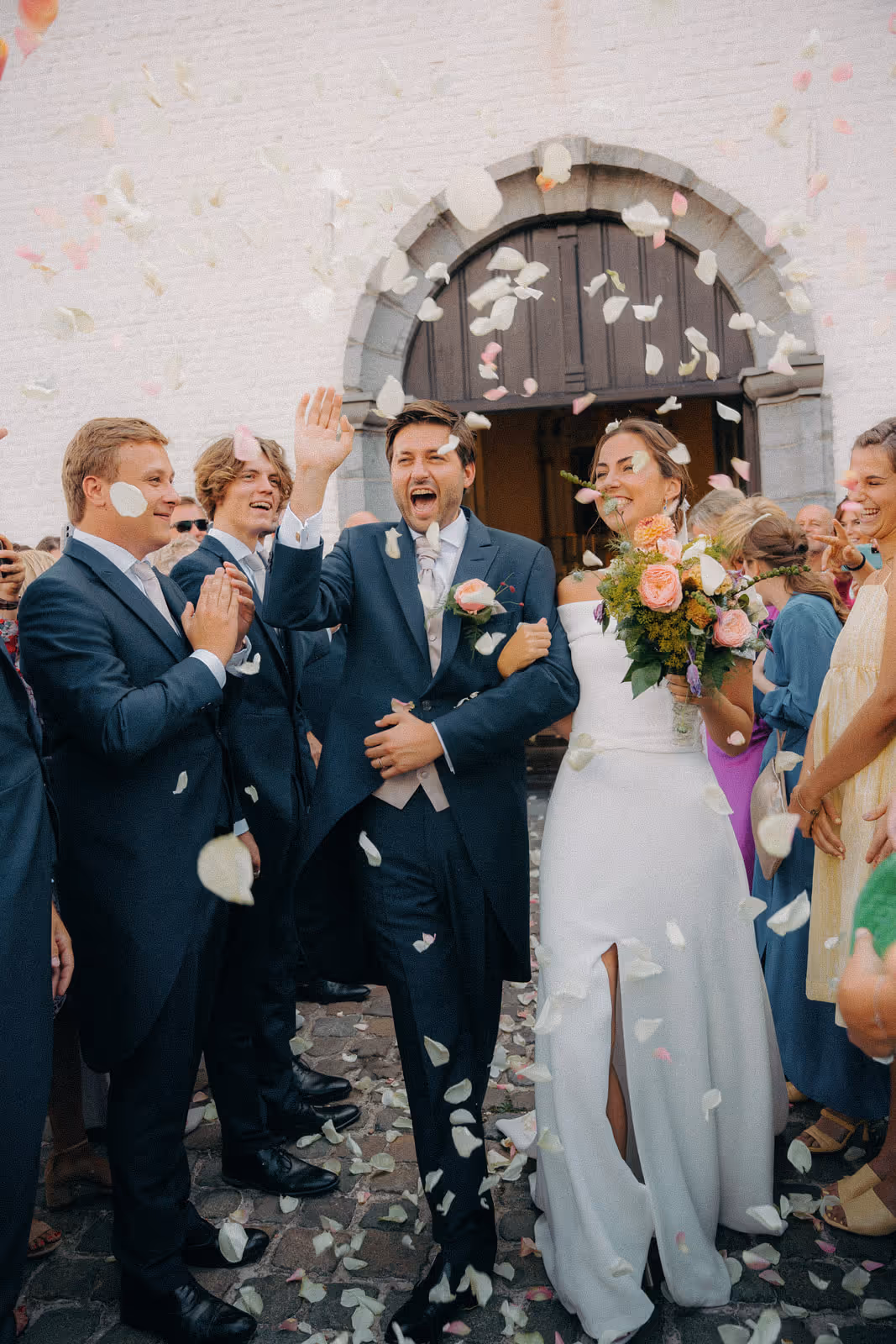 Bride and groom joyfully celebrating outside a church with flower petals being thrown and guests applauding.