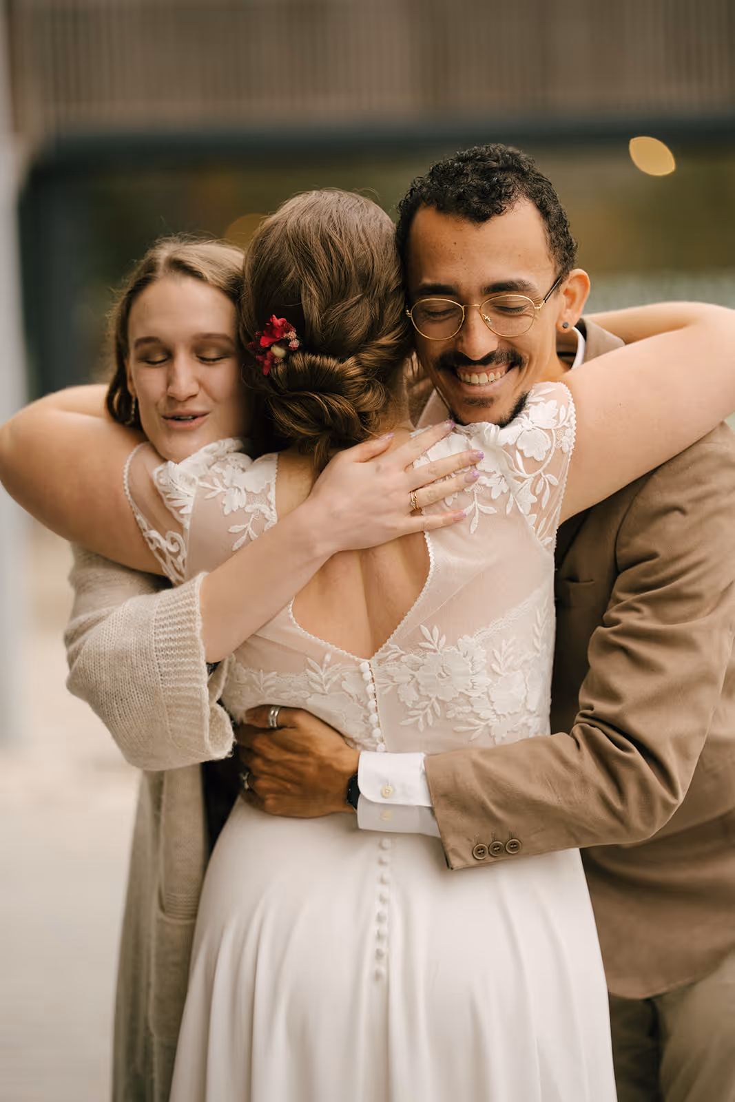 Smiling man and woman warmly hugging a bride in a white lace wedding dress with a floral hair accessory.