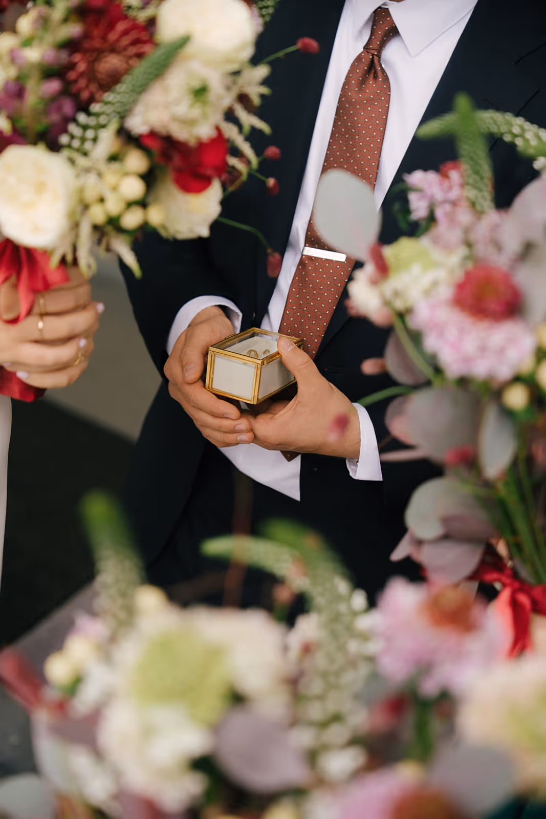 Person in a suit holding a small box with wedding rings, surrounded by blurred floral bouquets.