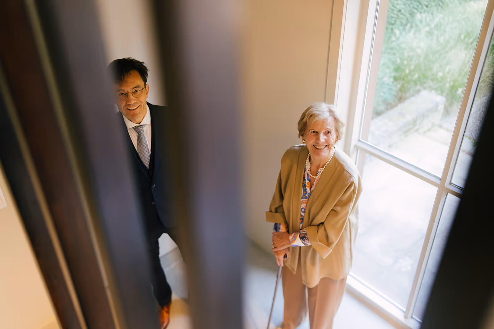 Smiling elderly woman with a cane and a man in a suit standing near a large window inside a bright room.