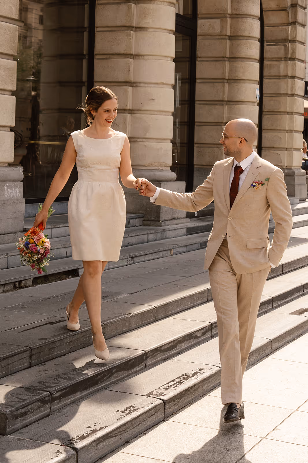 Bride and groom holding hands and walking down stone steps outside a building with columns.