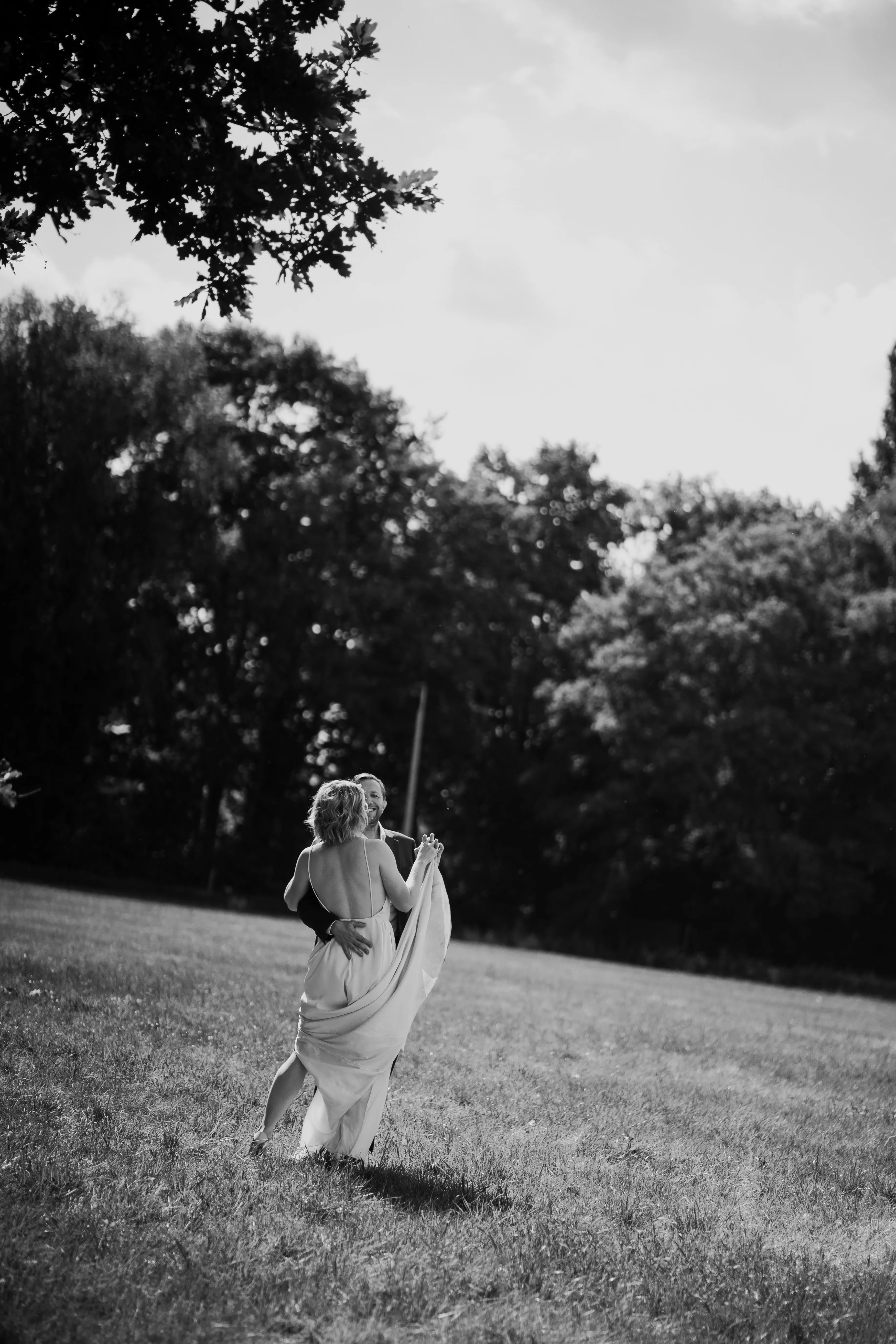 Black and white photo of a couple dancing in a grassy field with trees in the background.