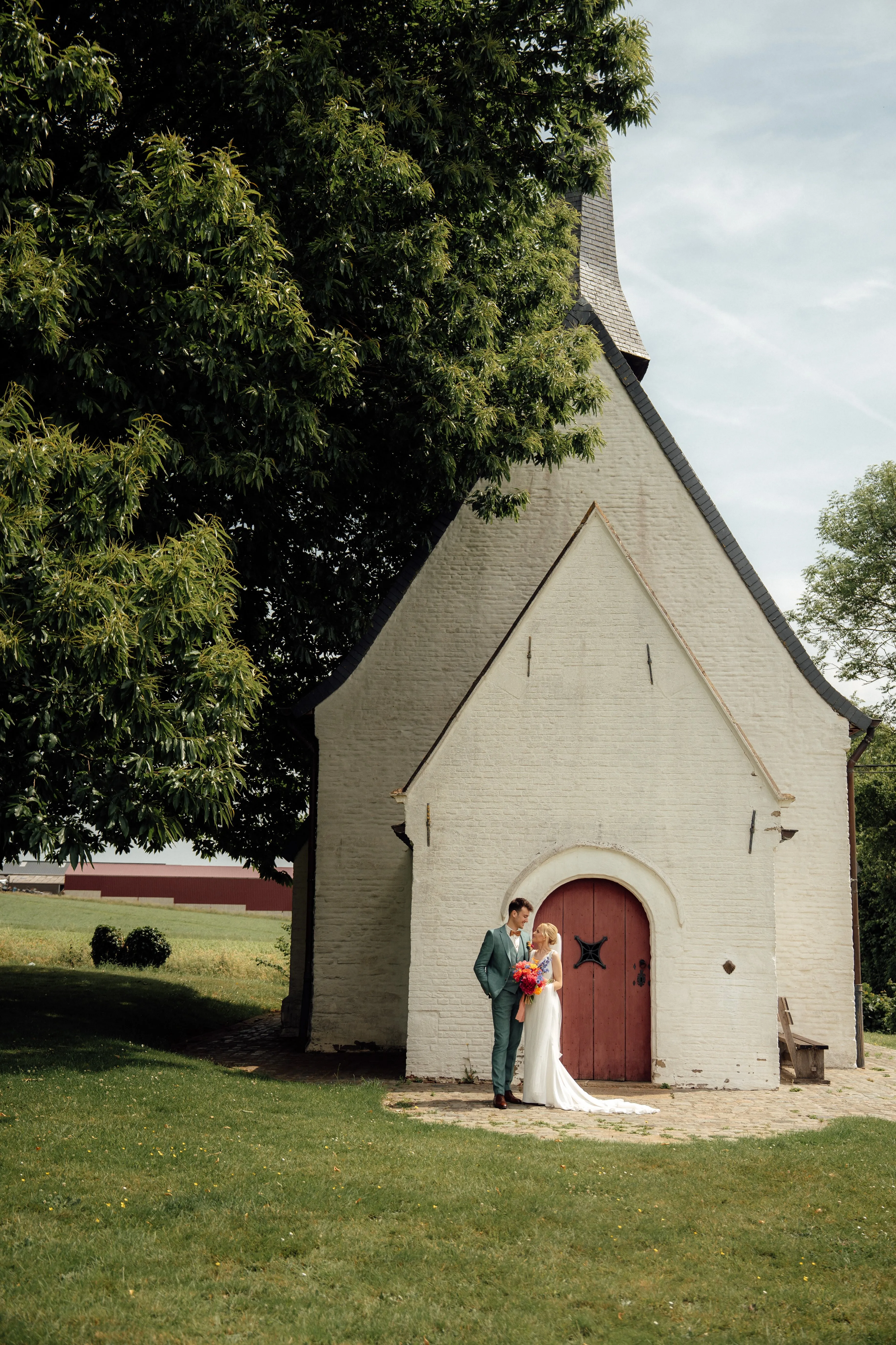 Bride in white gown holding a colorful bouquet and groom in green suit standing in front of a small white chapel with a red door under a large tree.