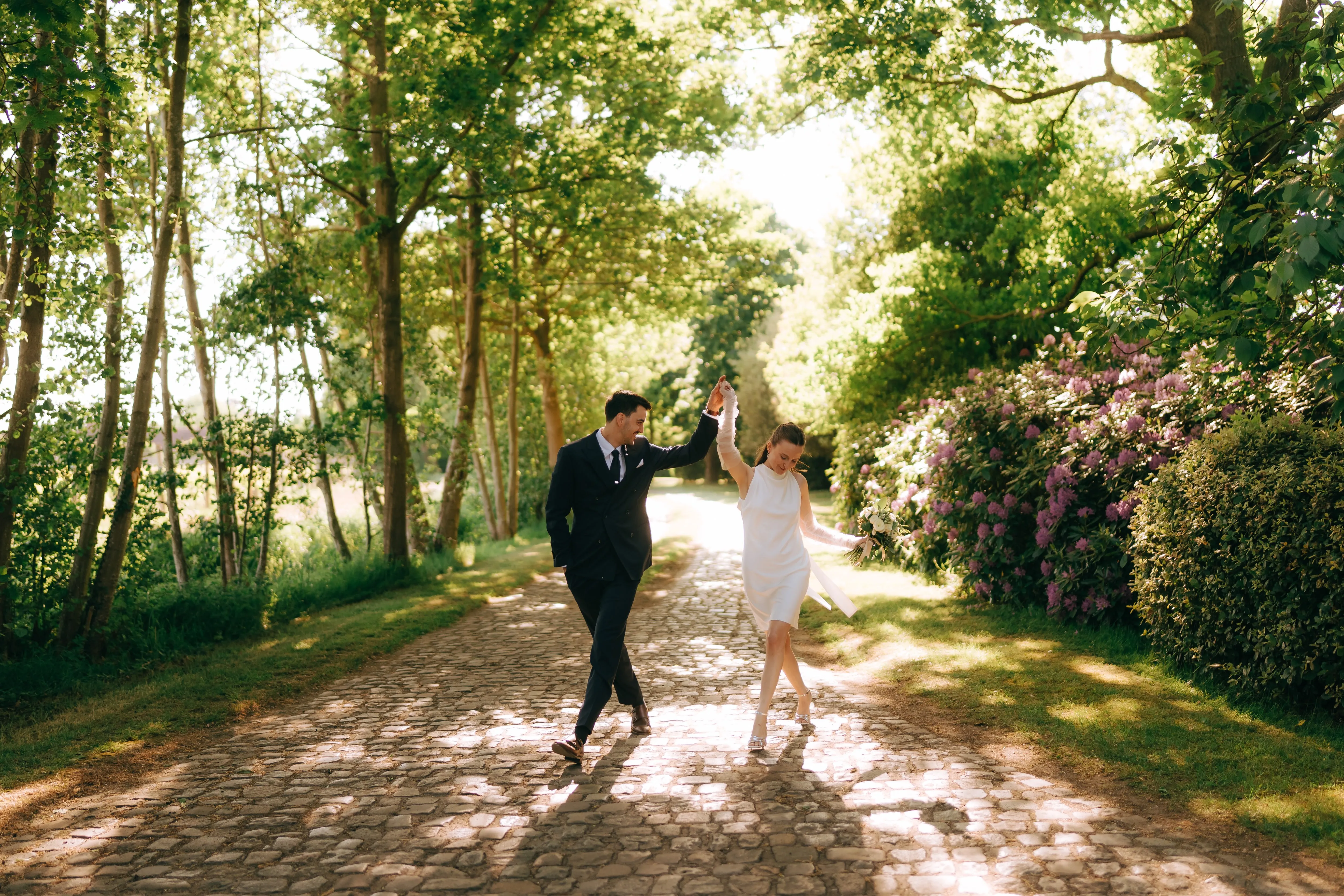 Bride and groom dancing on a sunlit cobblestone path surrounded by lush green trees and purple flowers.