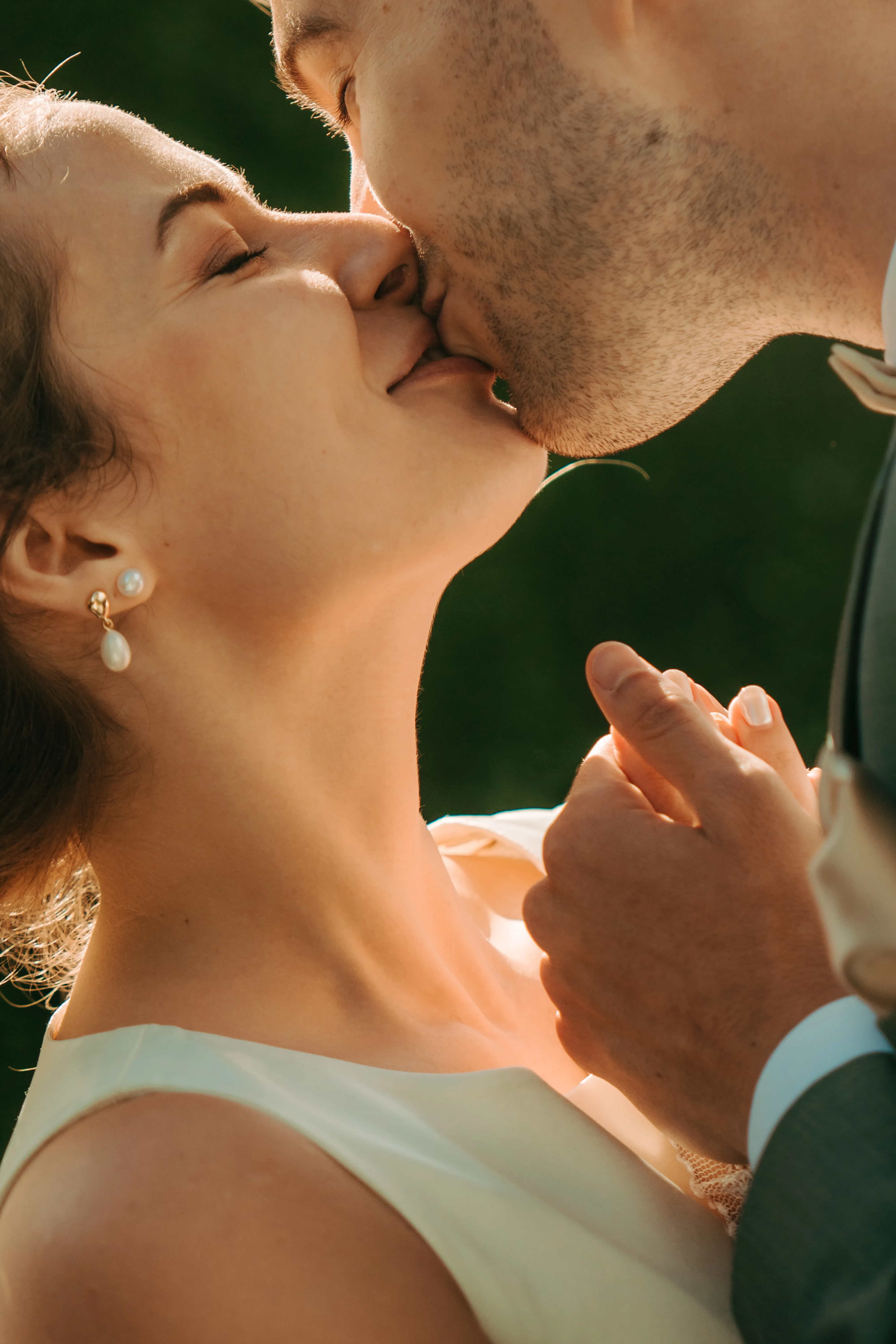 Close-up of a bride and groom gently kissing while holding hands outdoors.