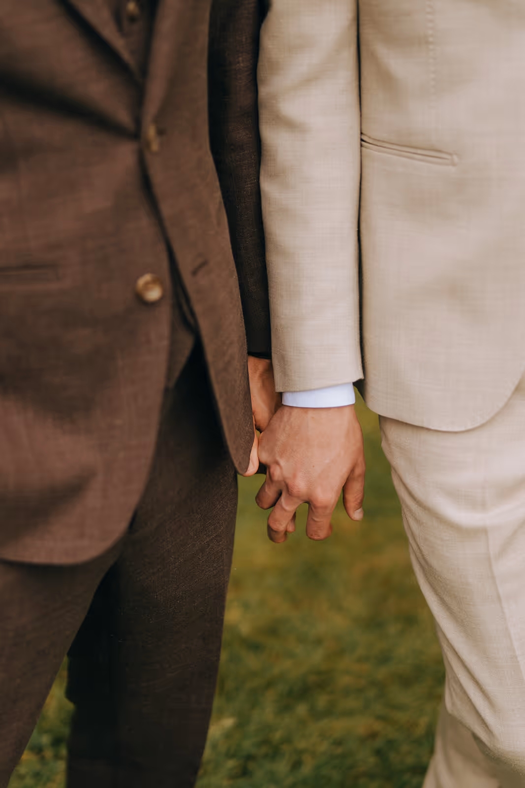 Two people dressed in suits holding hands outdoors on grass.