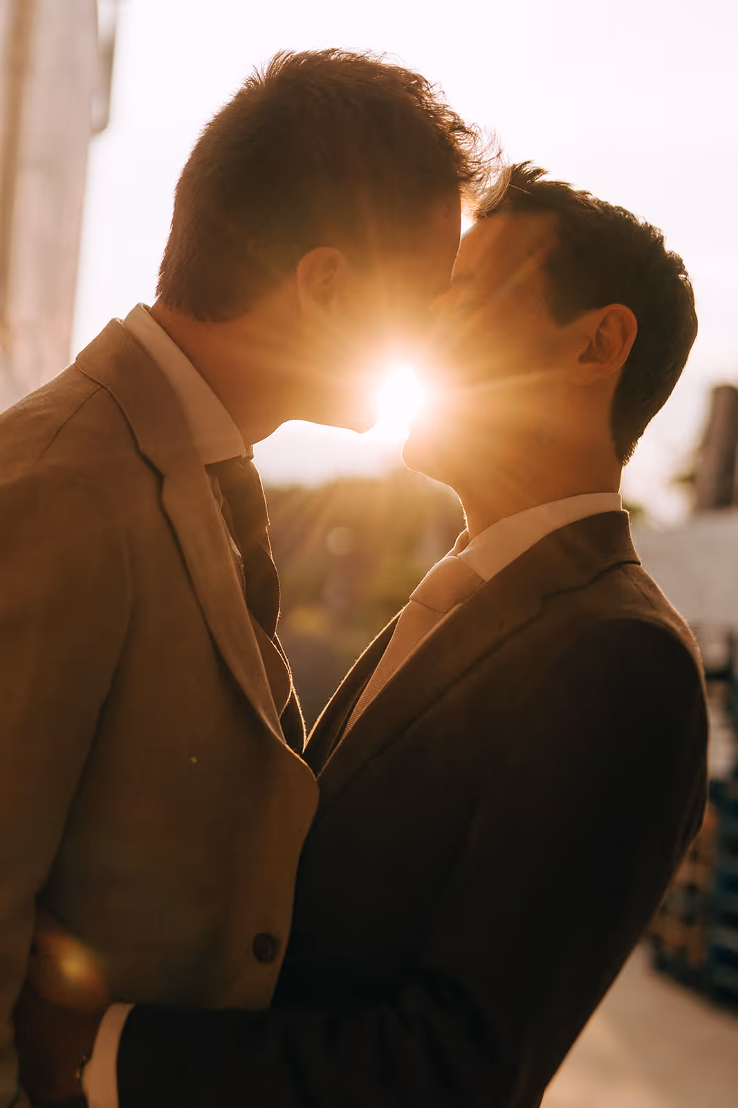Two men in suits sharing a kiss with sunlight shining between their faces in an outdoor setting.