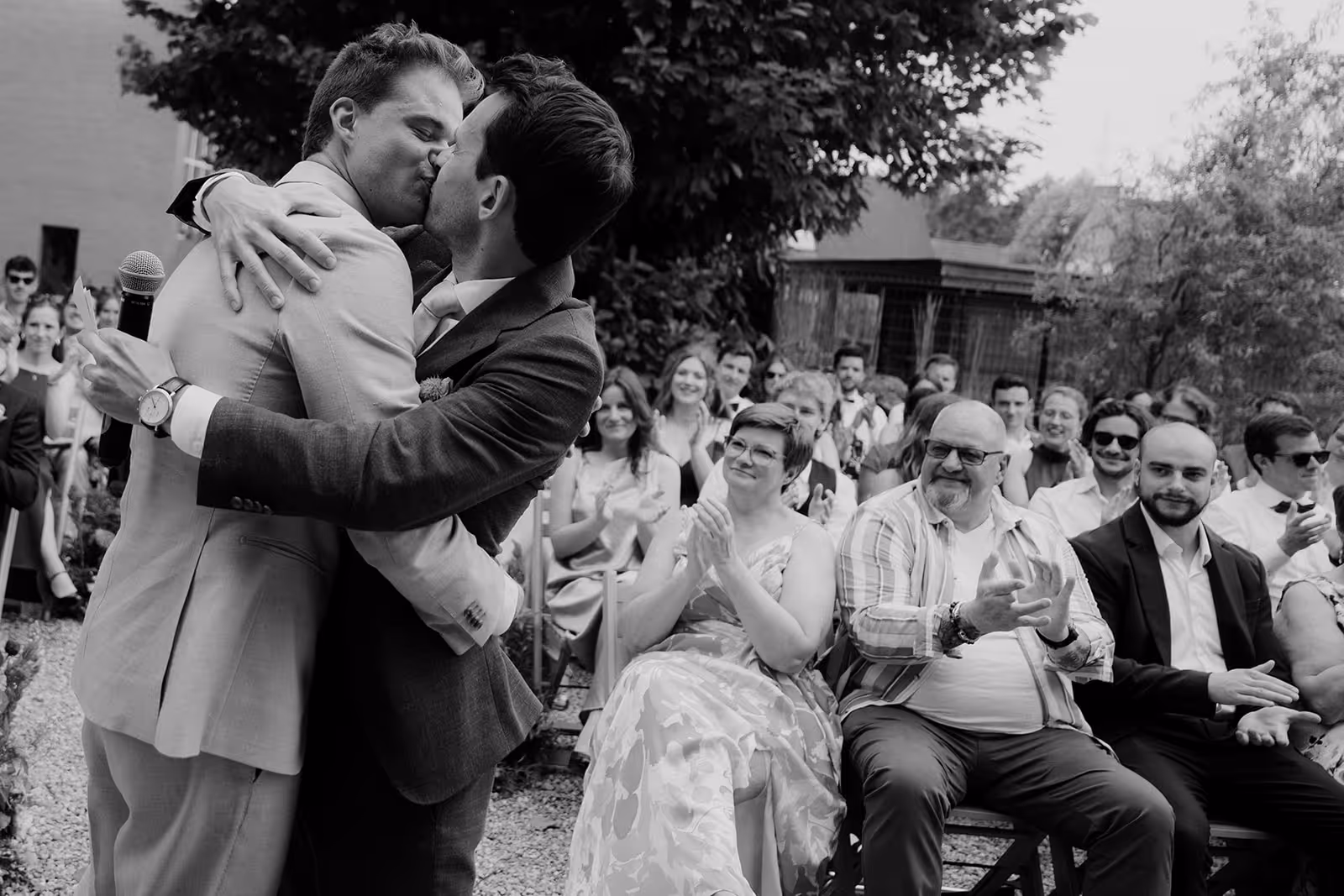 Two men in suits kissing and embracing at an outdoor wedding ceremony with seated guests clapping behind them.
