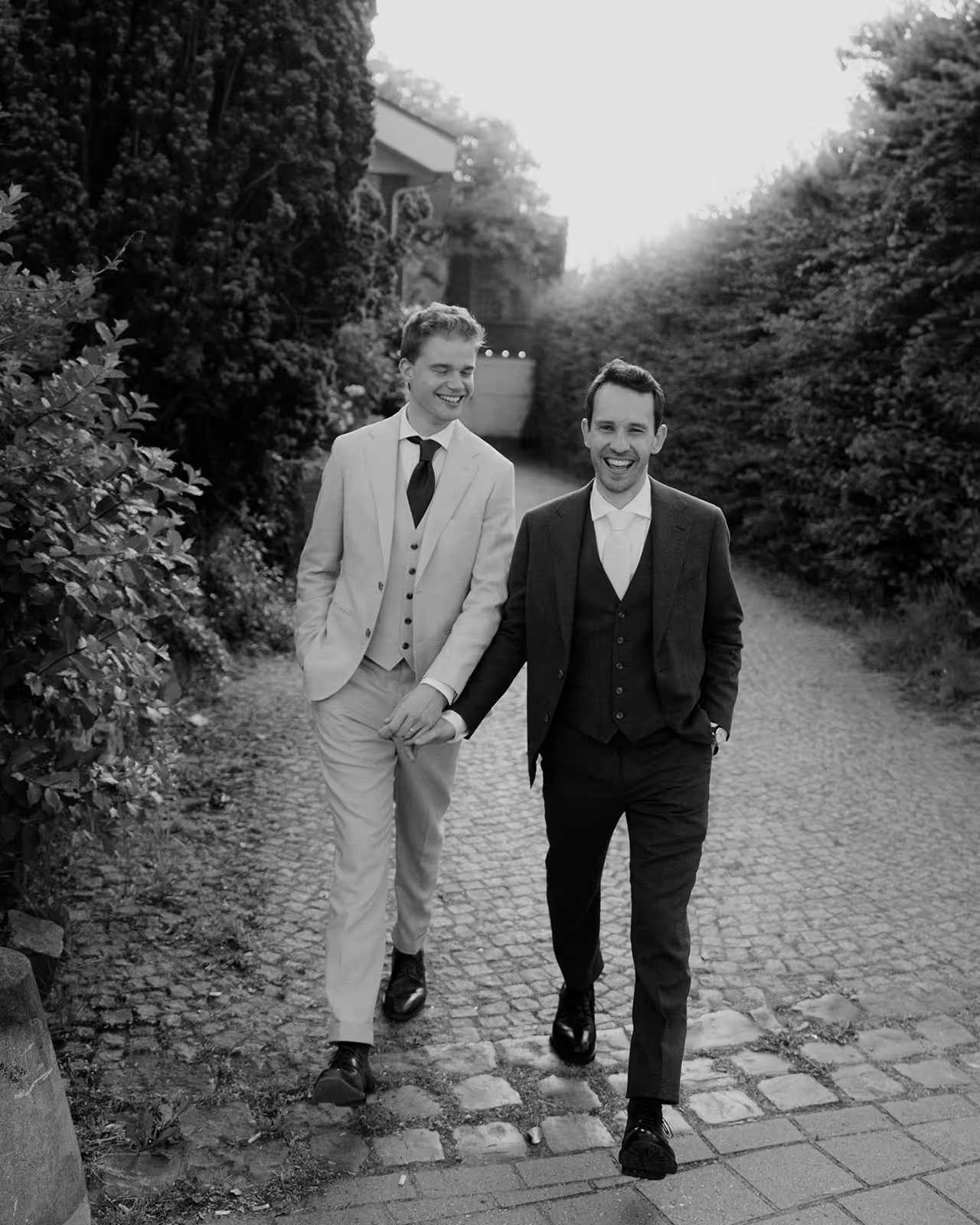 Two men in suits walking hand in hand down a cobblestone path, smiling happily.