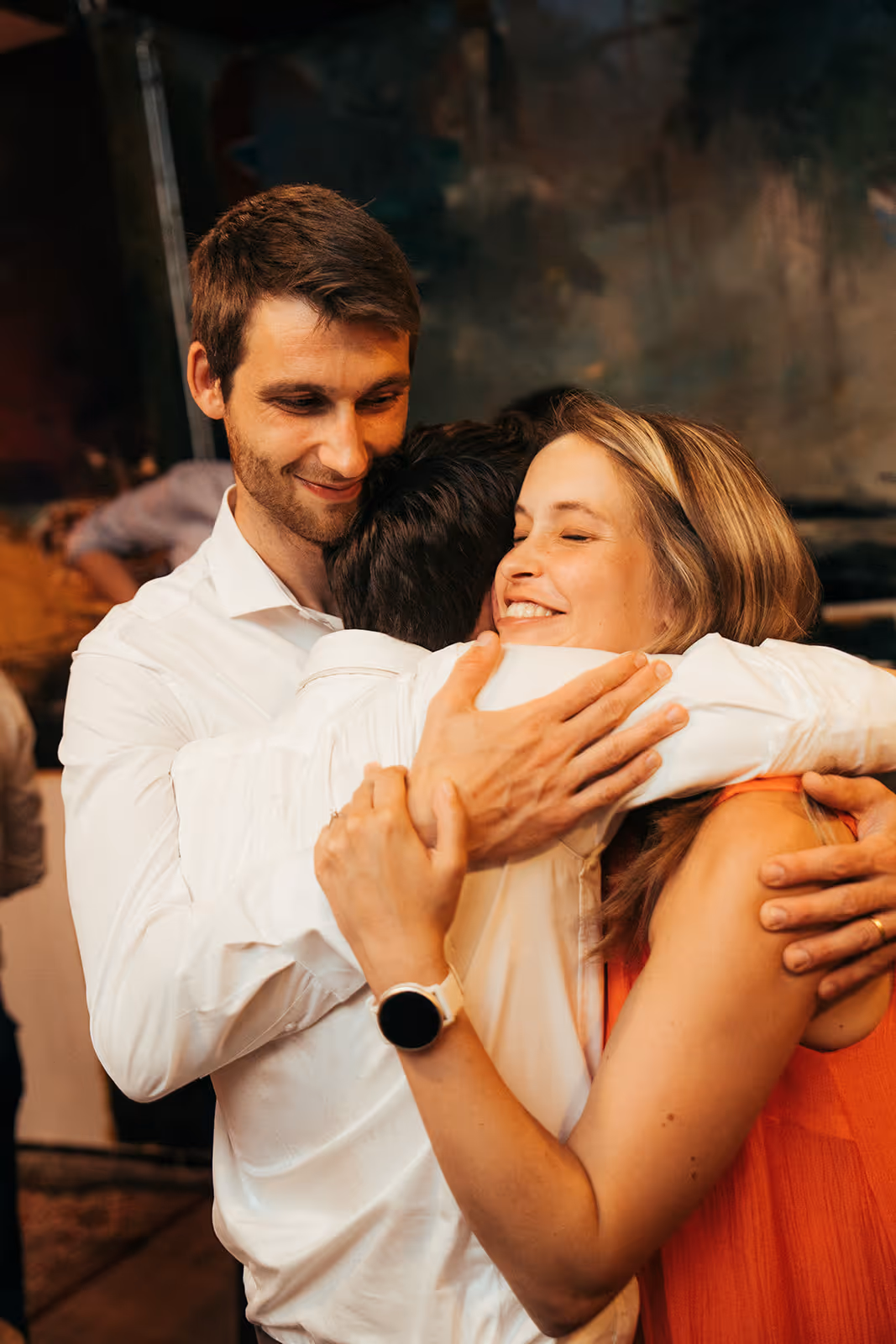 Three people warmly hugging each other, two wearing white shirts and one wearing an orange sleeveless top, smiling with closed eyes.