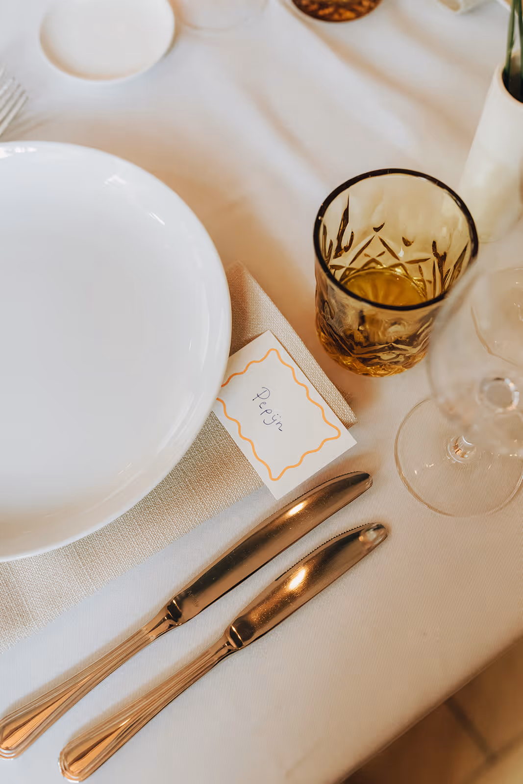 Table setting with two knives, a white plate, an amber glass with liquid, a wine glass, and a name card labeled 'Pepijn' on a beige napkin.