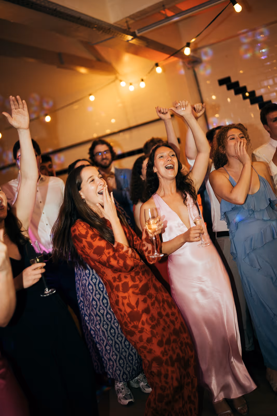 Group of people dancing and celebrating indoors with drinks and festive string lights overhead.