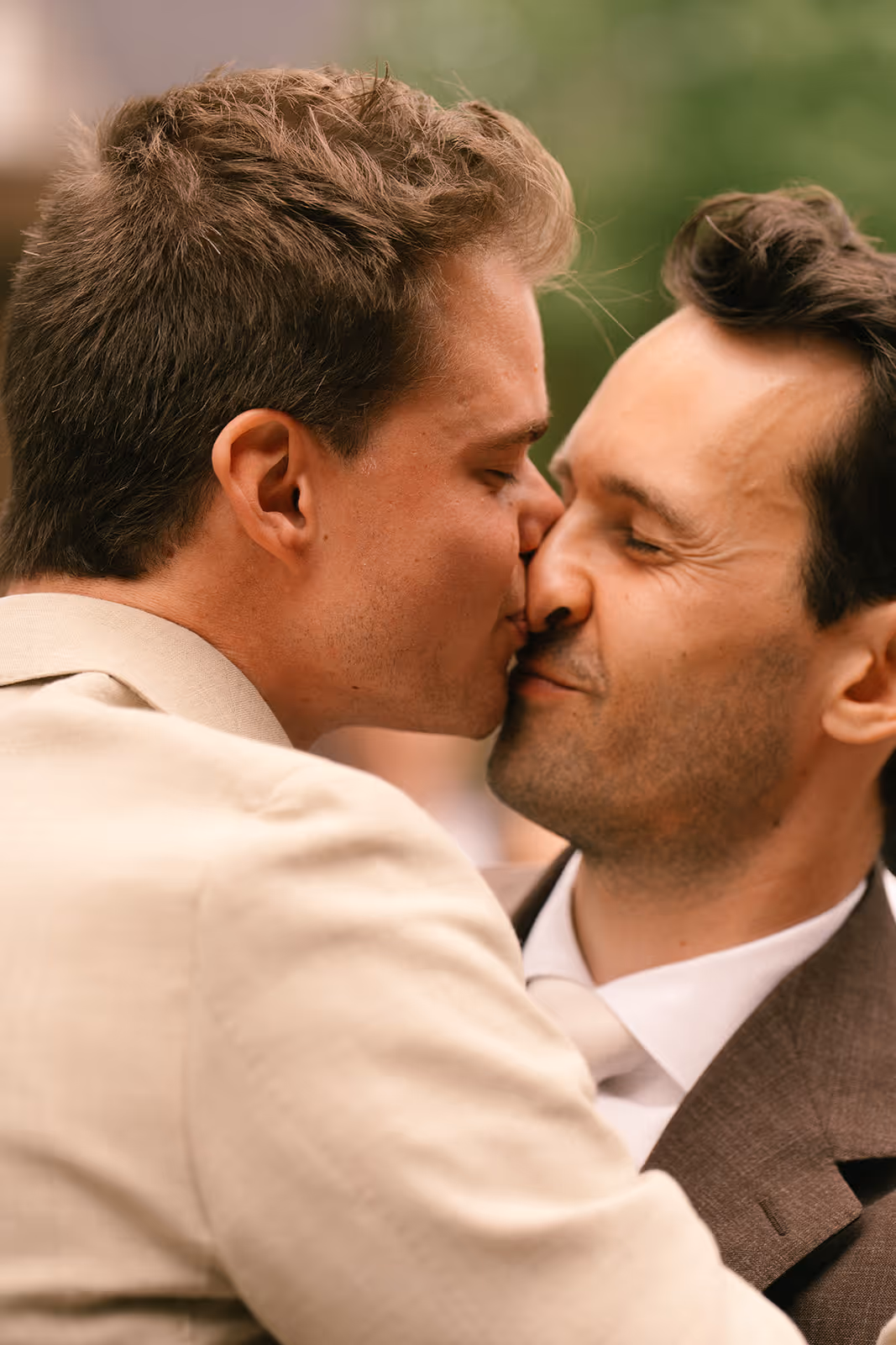 Two men in suits sharing a close, affectionate kiss outdoors.