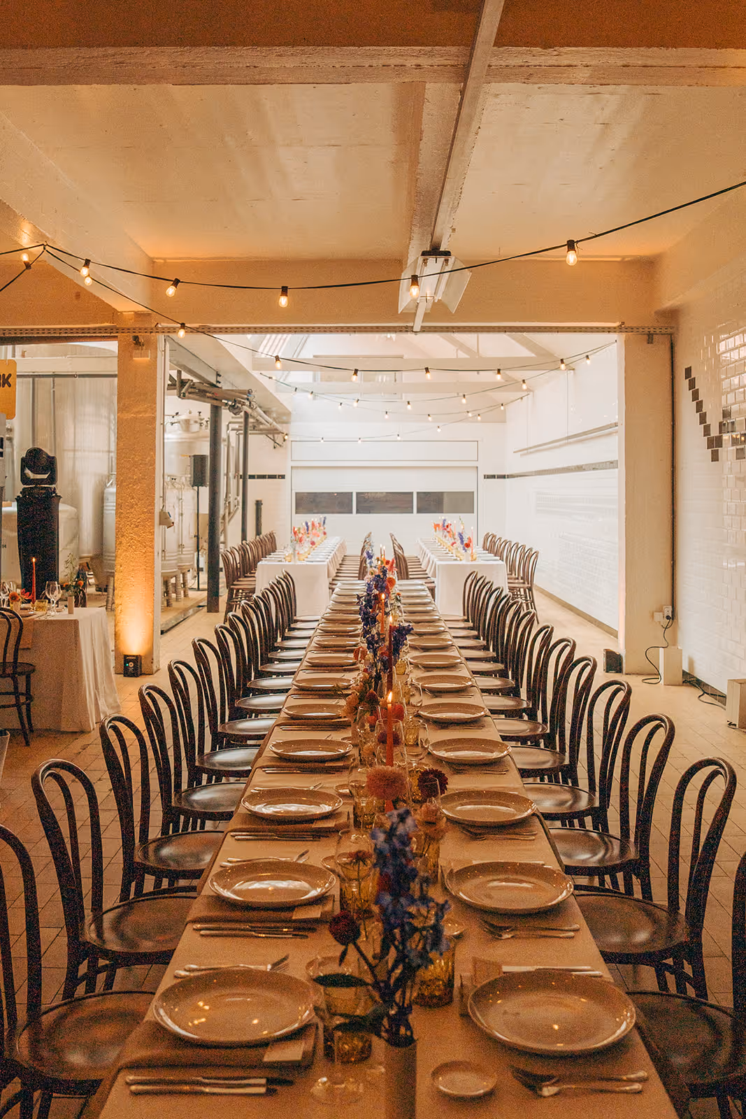 Long dining tables set for a formal event with white plates, silver cutlery, floral centerpieces, and string lights overhead in an industrial-style room.