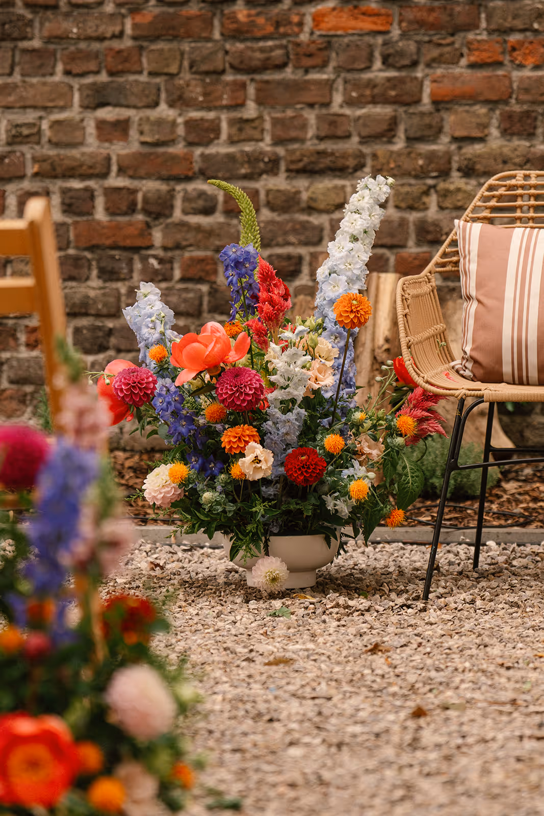 Colorful floral arrangement with red, orange, blue, and white flowers in a white vase on gravel, next to a wicker chair with striped cushion and a brick wall background.